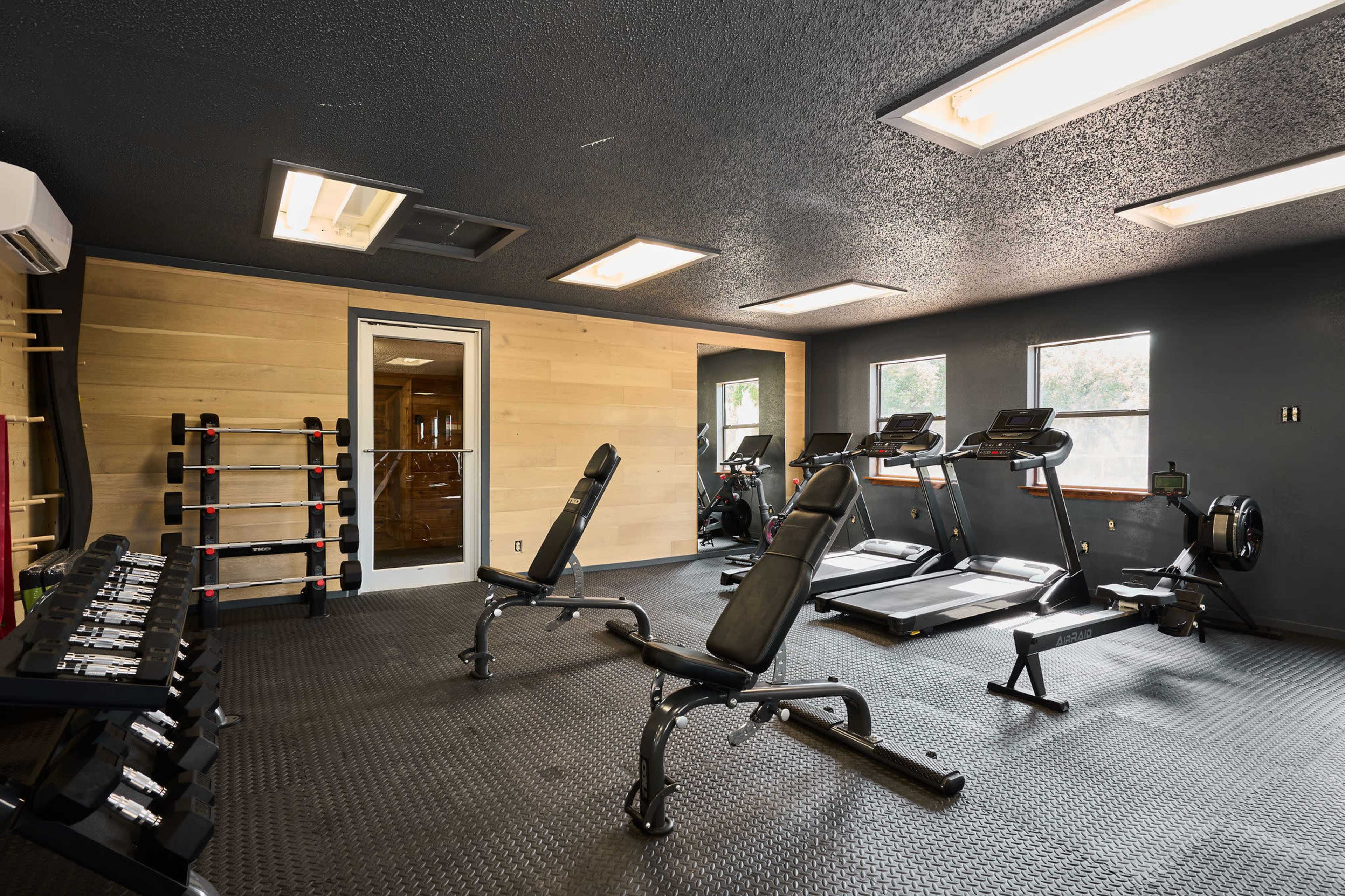The image shows a modern gym with exercise equipment, including treadmills, a rowing machine, dumbbells, and benches on a textured floor.