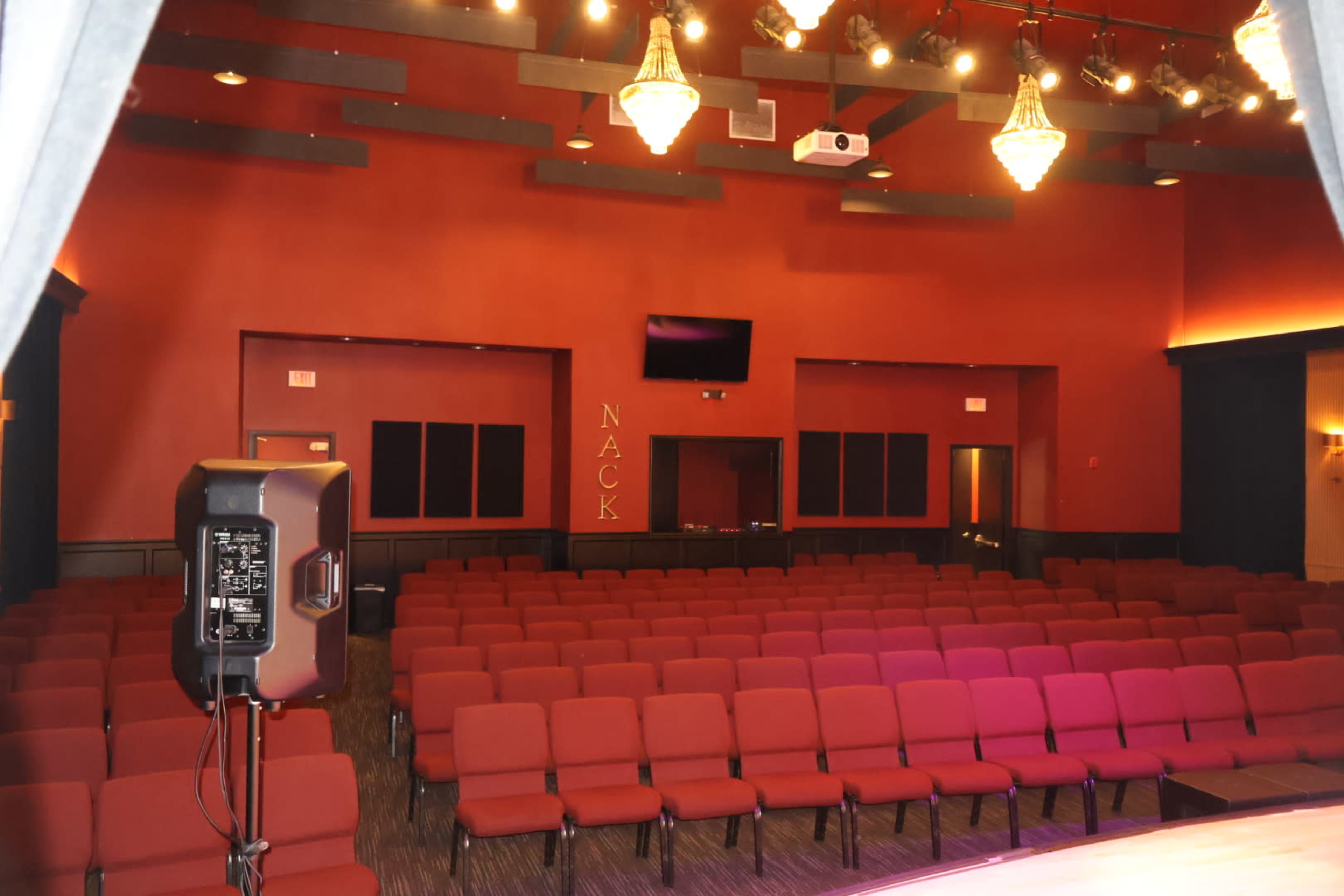 The image shows the interior of a theater with rows of red chairs facing a stage, illuminated by overhead lights.