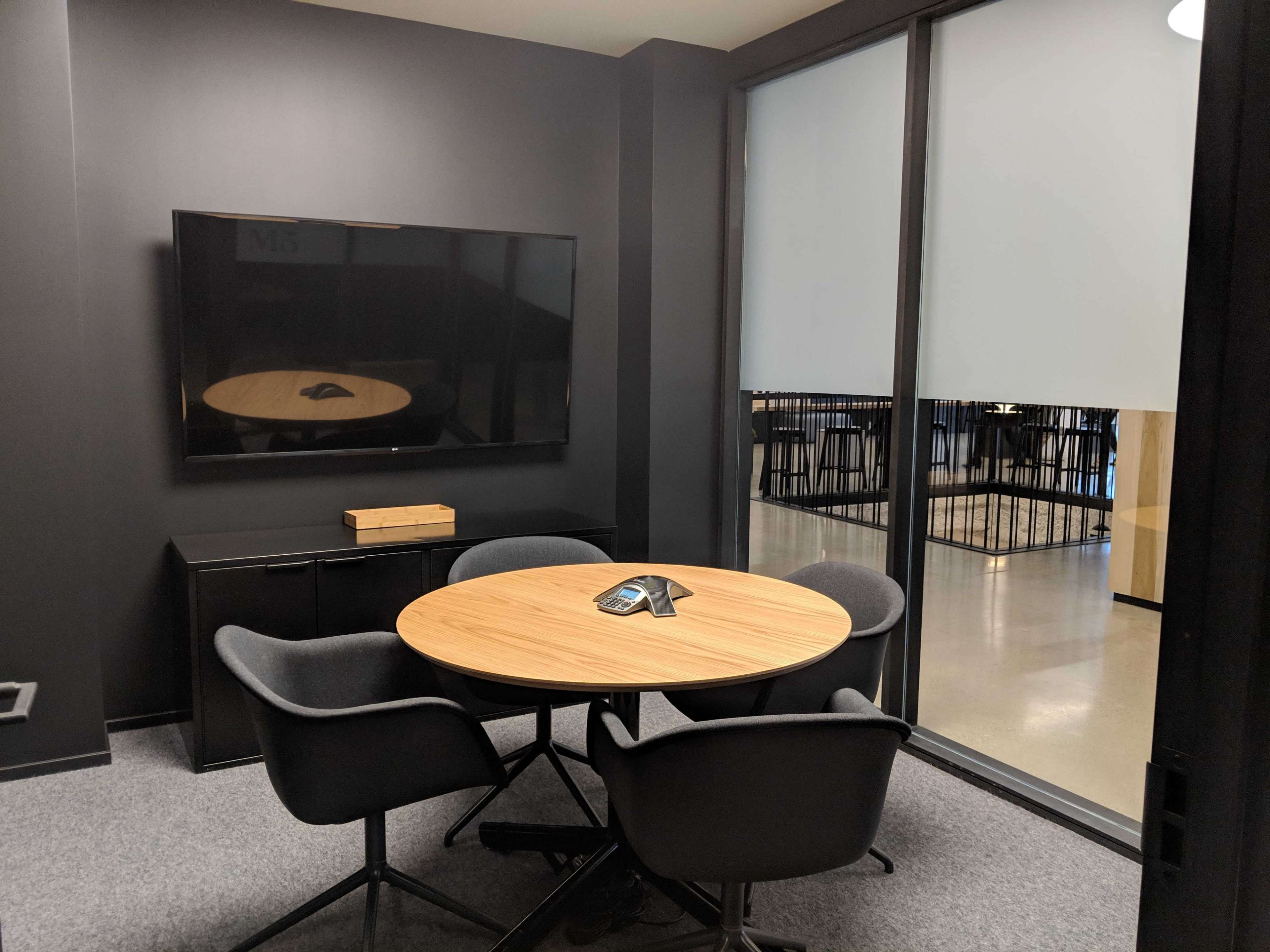 A small conference room features a round wooden table surrounded by four gray chairs, with a large screen mounted on the wall.