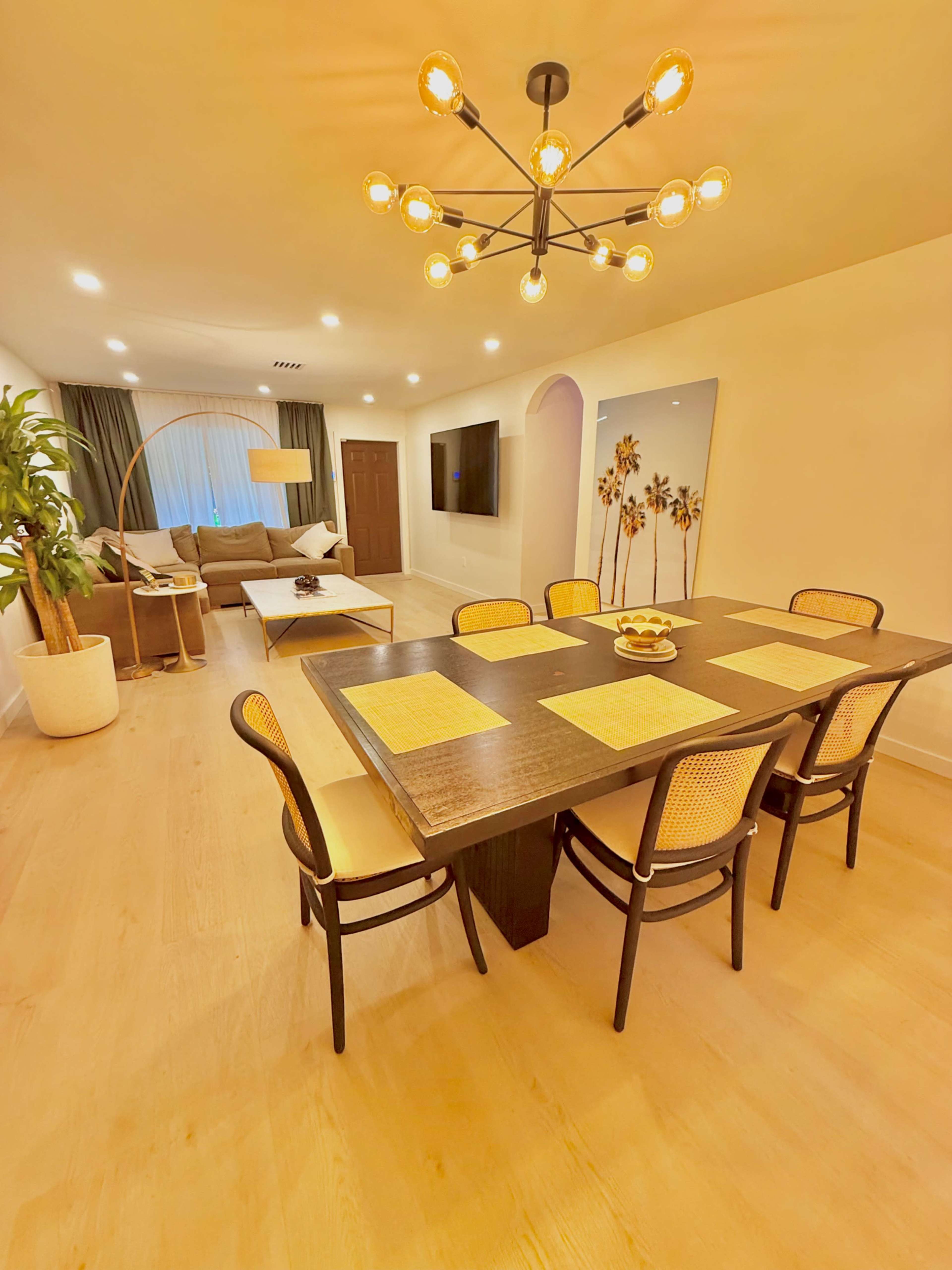 The image shows a modern dining area with a large table, six chairs, and a living space featuring a sofa, television, and decorative elements.