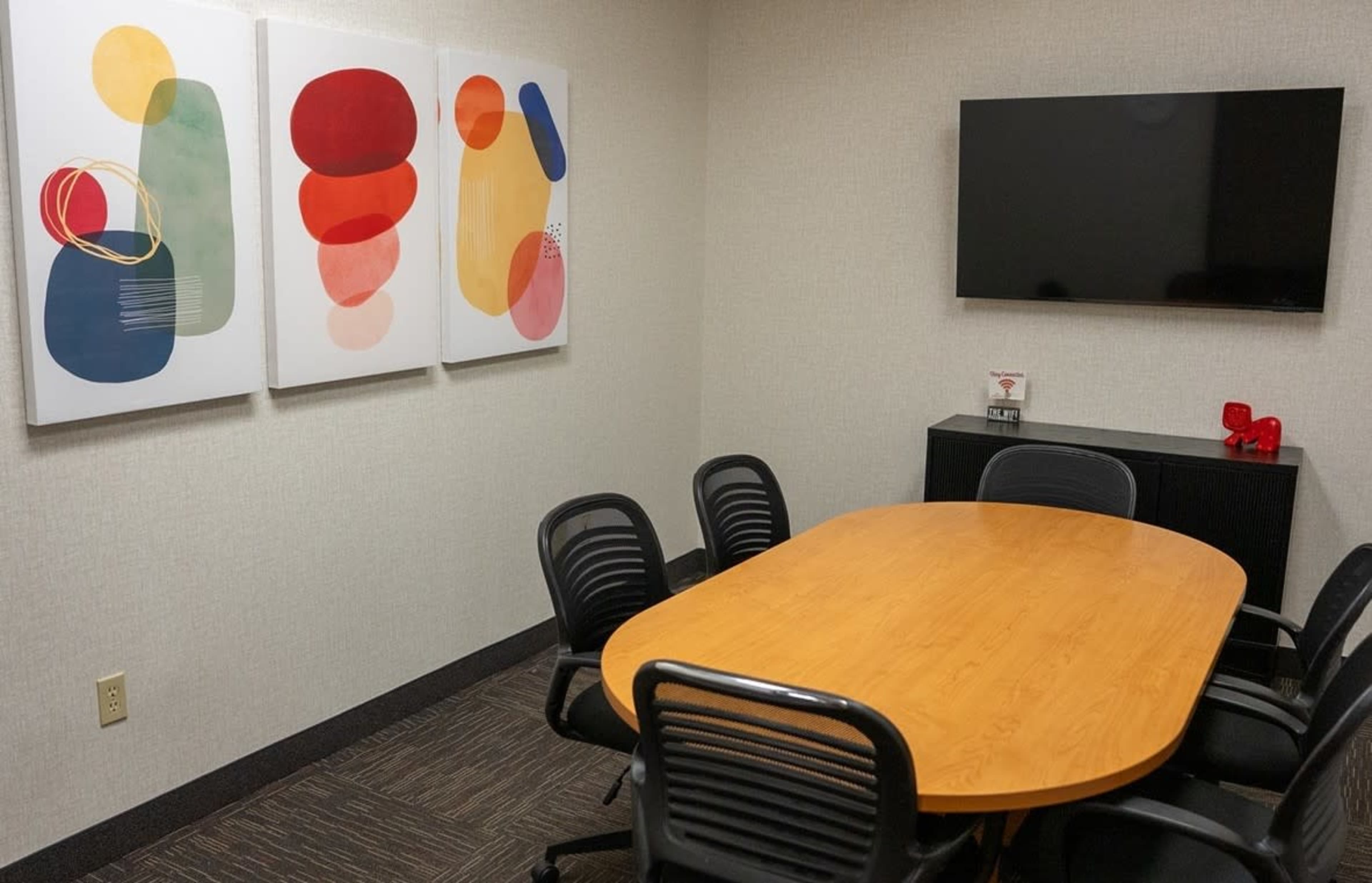 The image shows a meeting room with a round wooden table surrounded by black chairs, three abstract paintings on the wall, and a television mounted on the opposite wall.