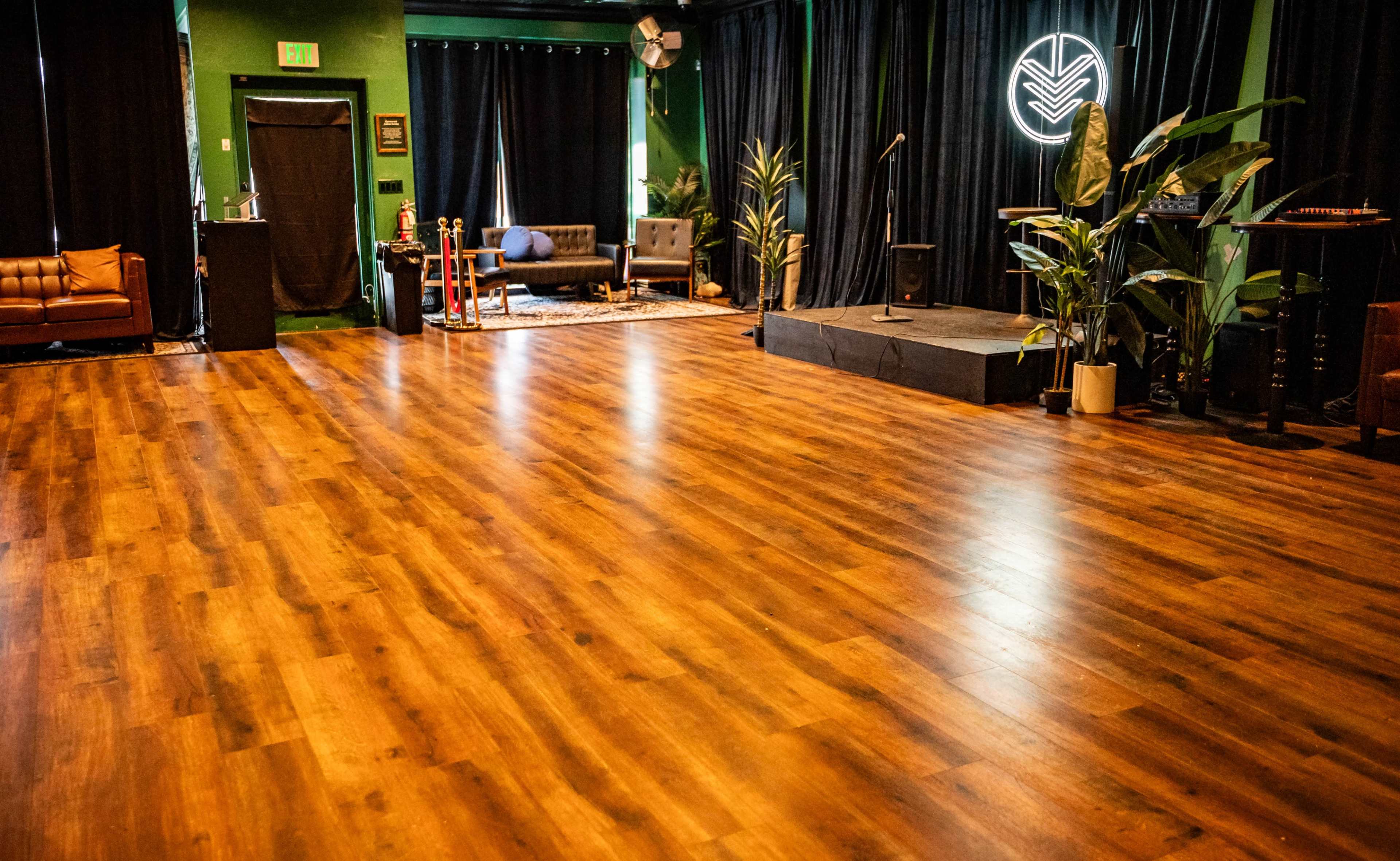 A performance space with a wooden floor, a small stage, seating areas, and plants along the walls.