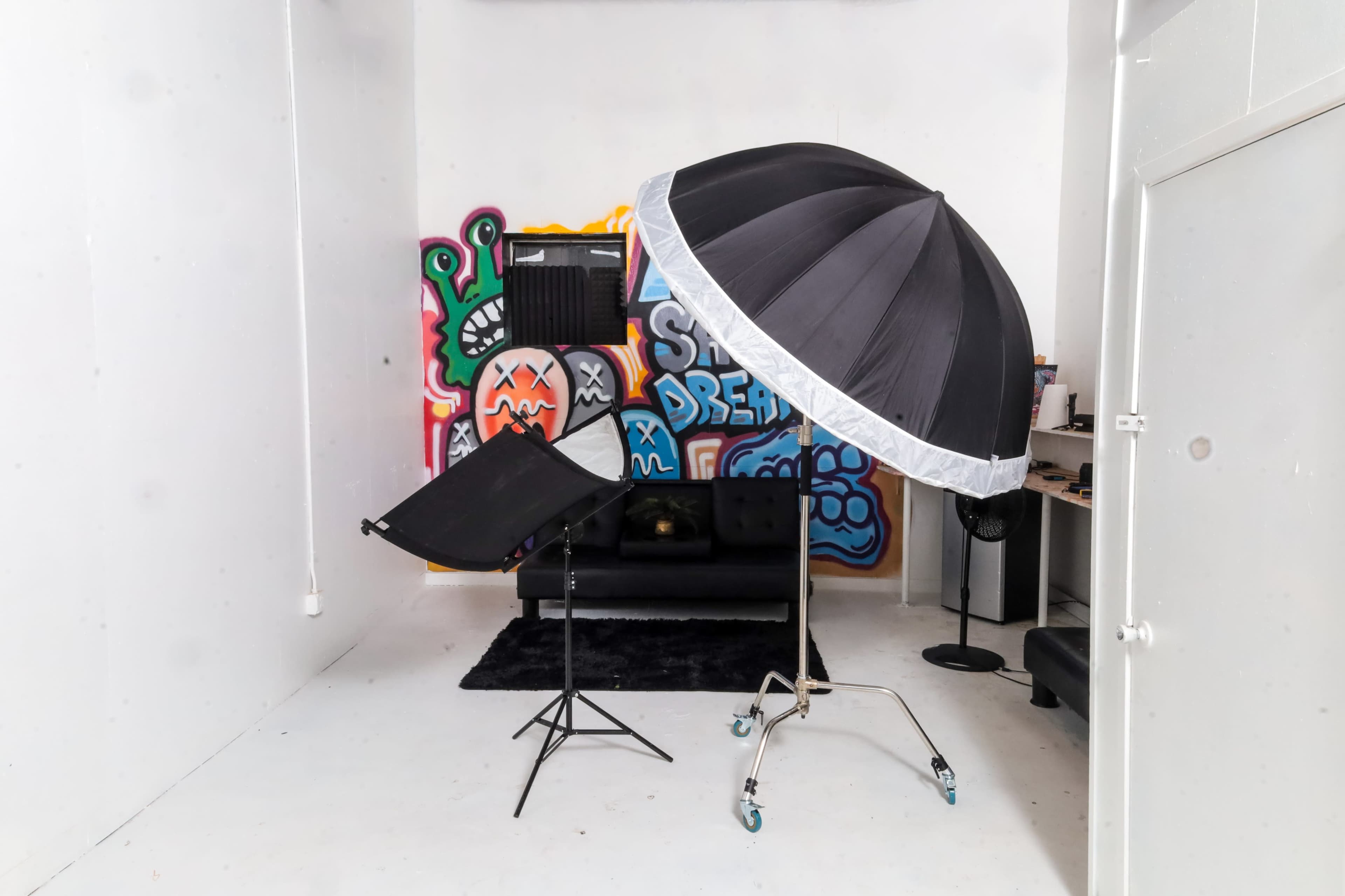 The image shows a photography studio with a large black umbrella light setup and a black couch against a colorful graffiti wall.