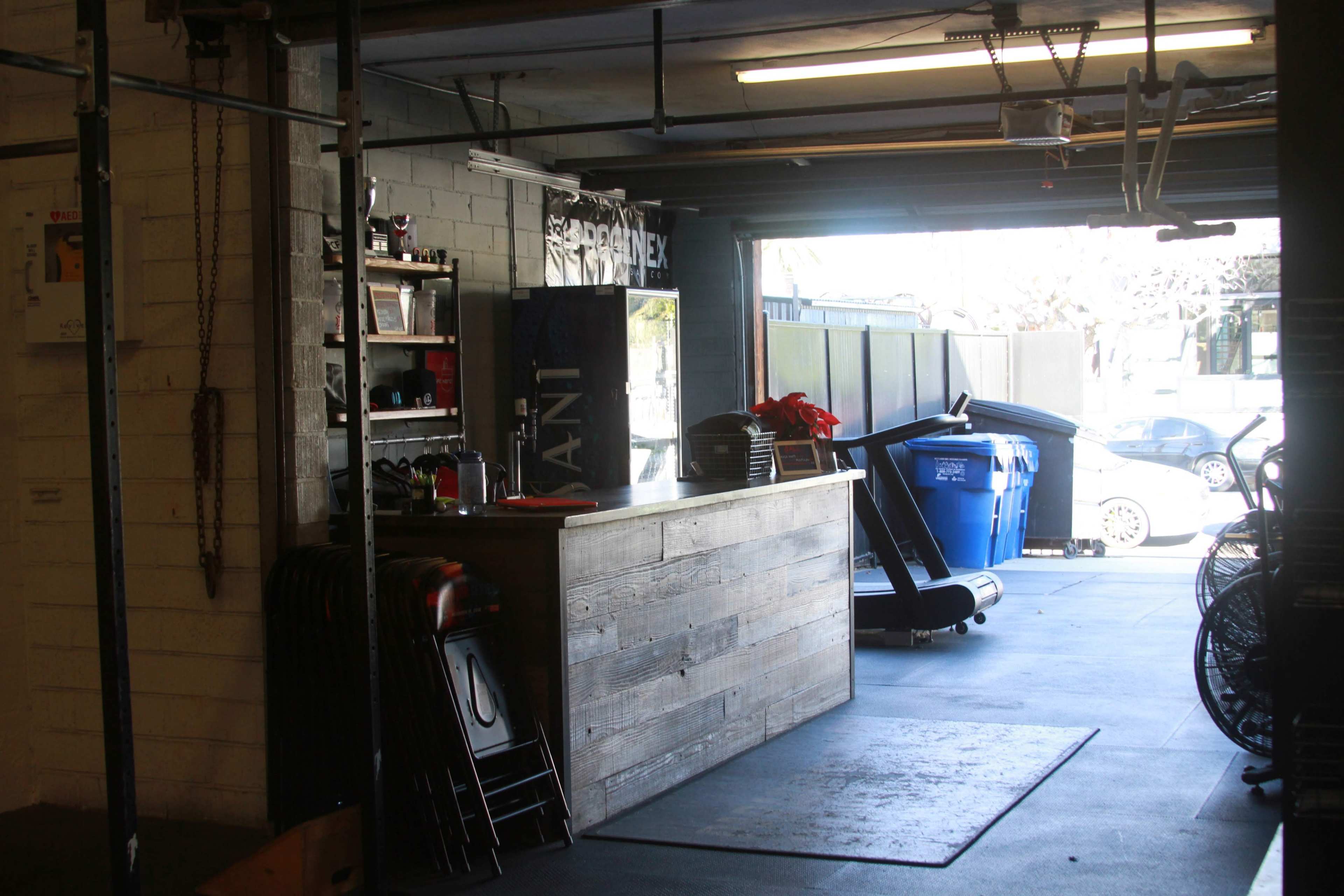 An interior space of a gym or fitness facility with a wooden reception counter, shelves with equipment, and a treadmill near a blue recycling bin.
