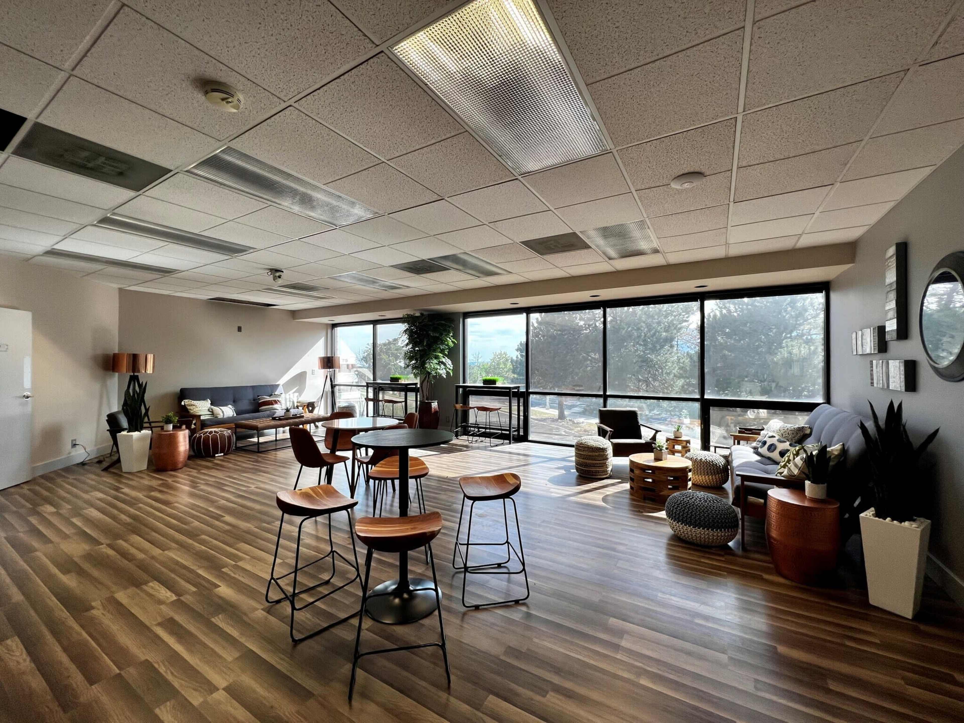 The image shows a spacious, well-lit common area with modern furniture, large windows, and a wooden floor.
