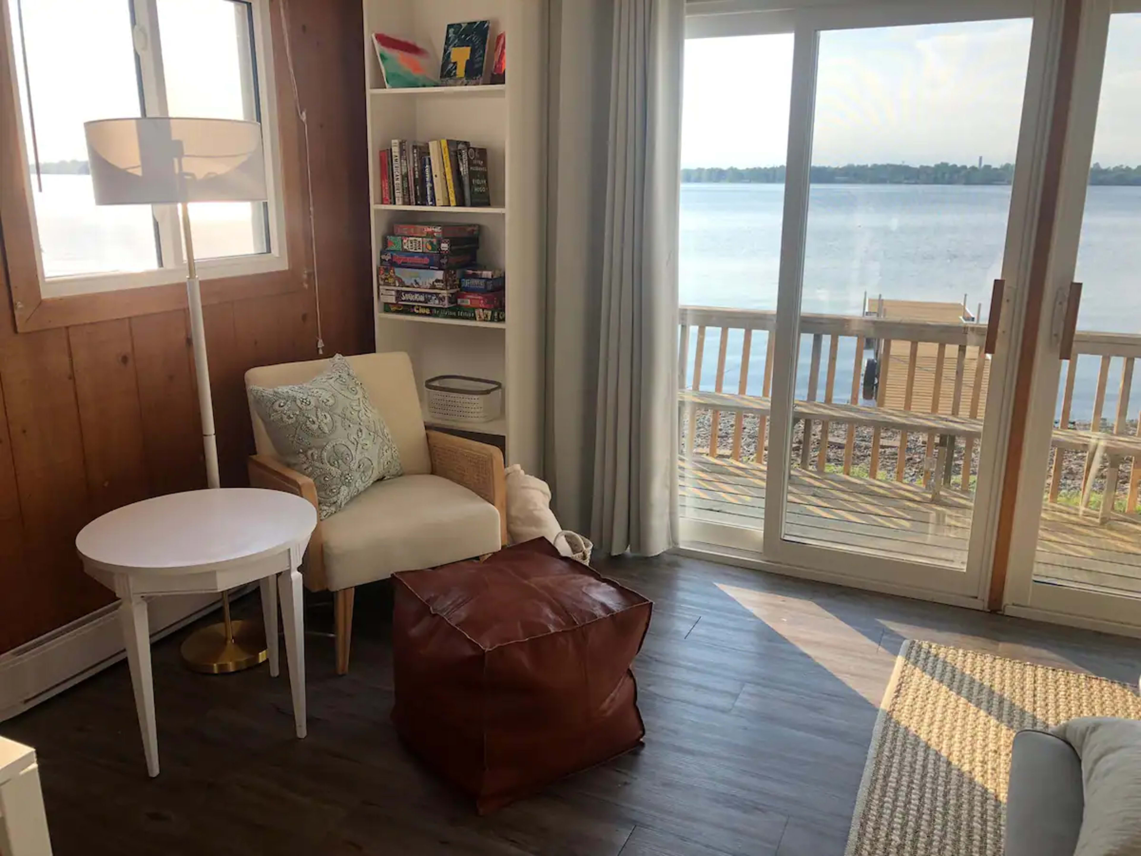 A cozy corner of a room features a light-colored armchair, a round white table, a brown pouf, and a large window overlooking a calm lake.