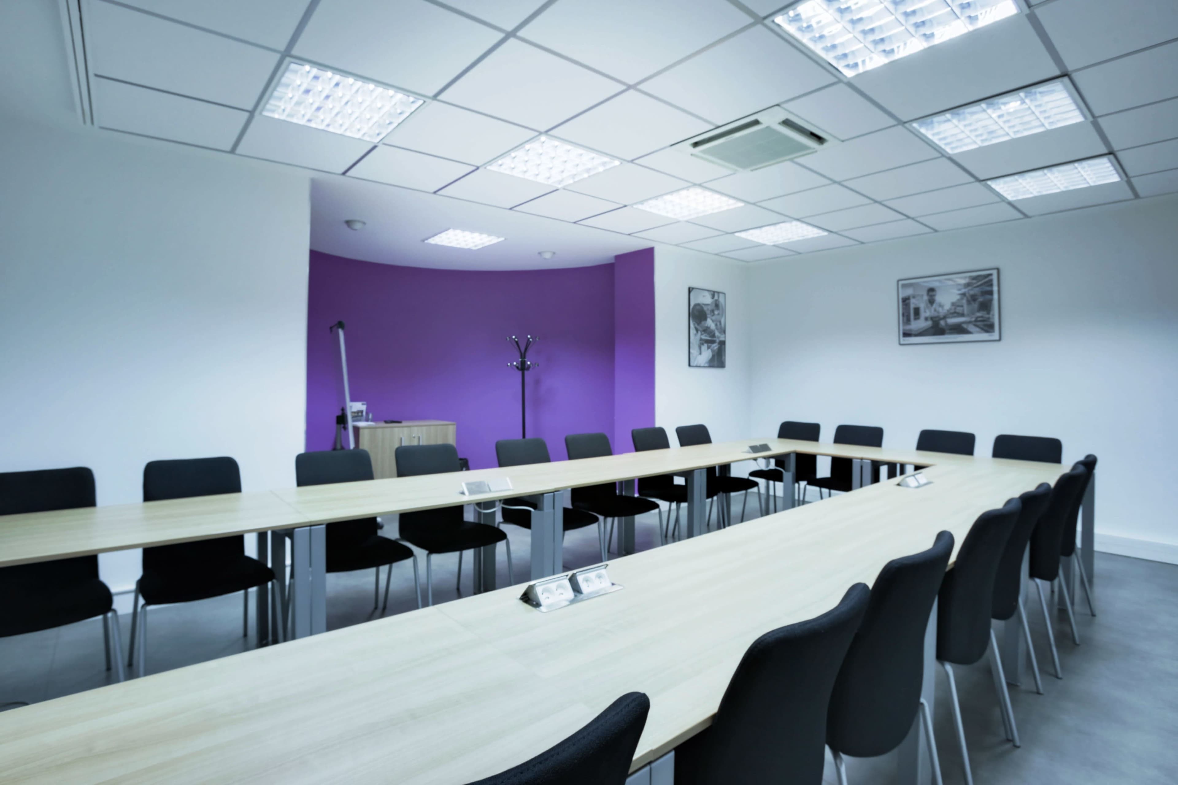 A modern conference room features a long wooden table surrounded by black chairs, with a purple accent wall and overhead lighting.