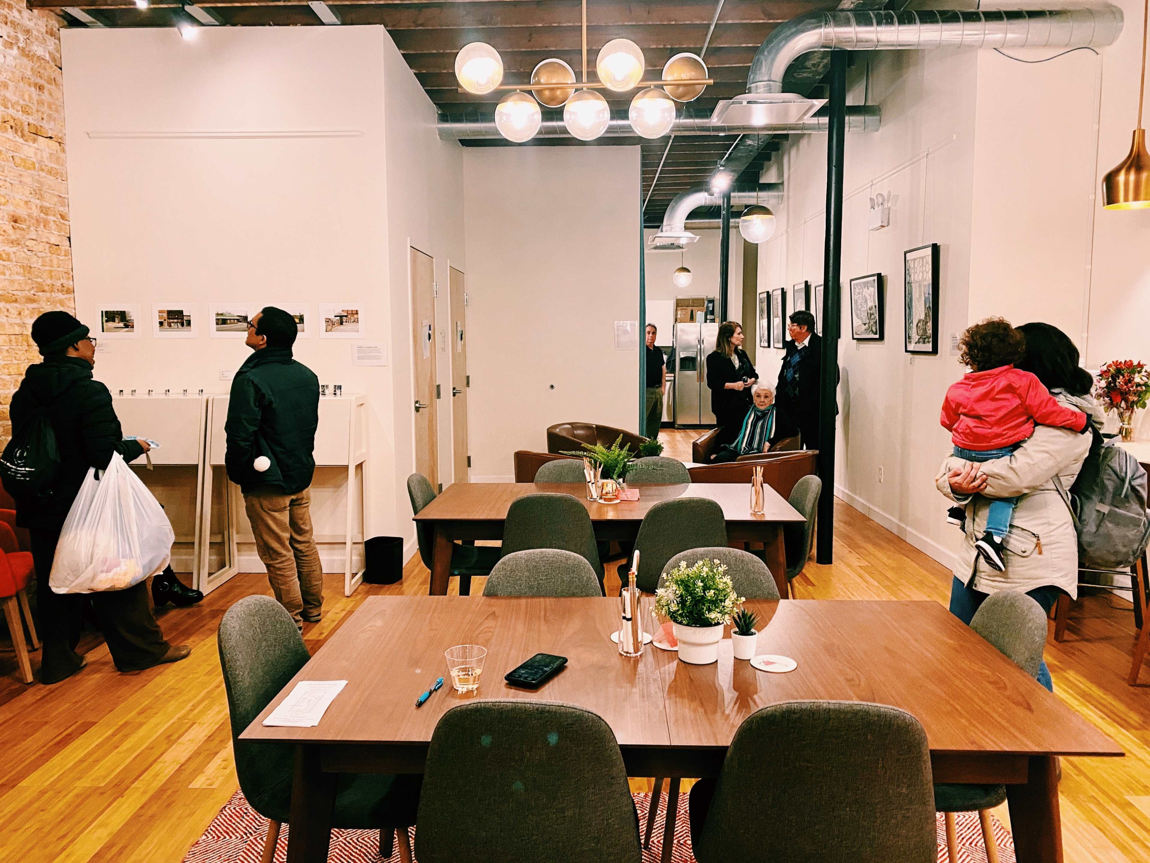 A spacious interior with wooden tables and chairs, where several people are engaged in various activities, including browsing a wall display and conversing.