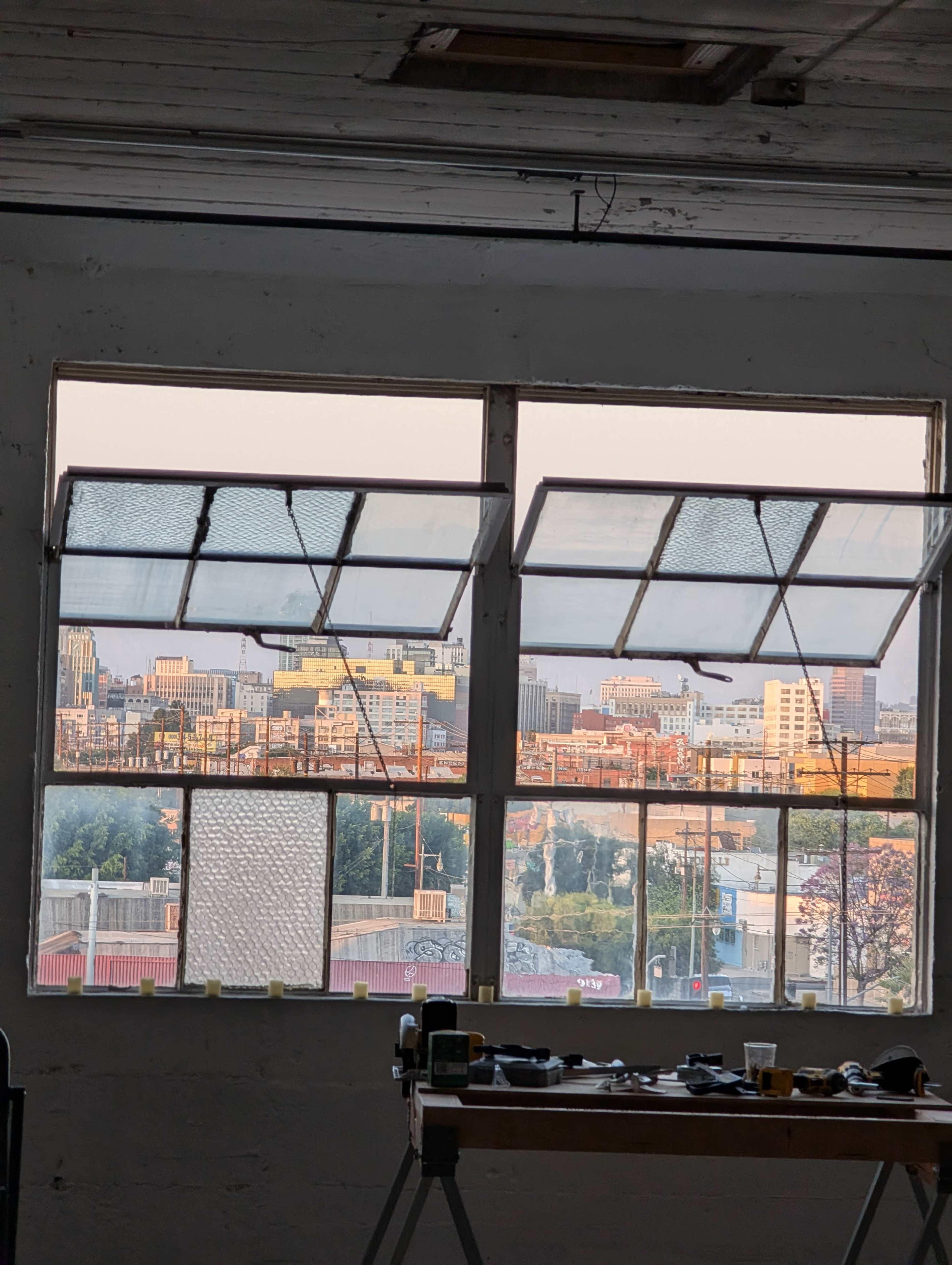 The image shows a workshop interior with a large window offering a view of a city skyline at sunset.