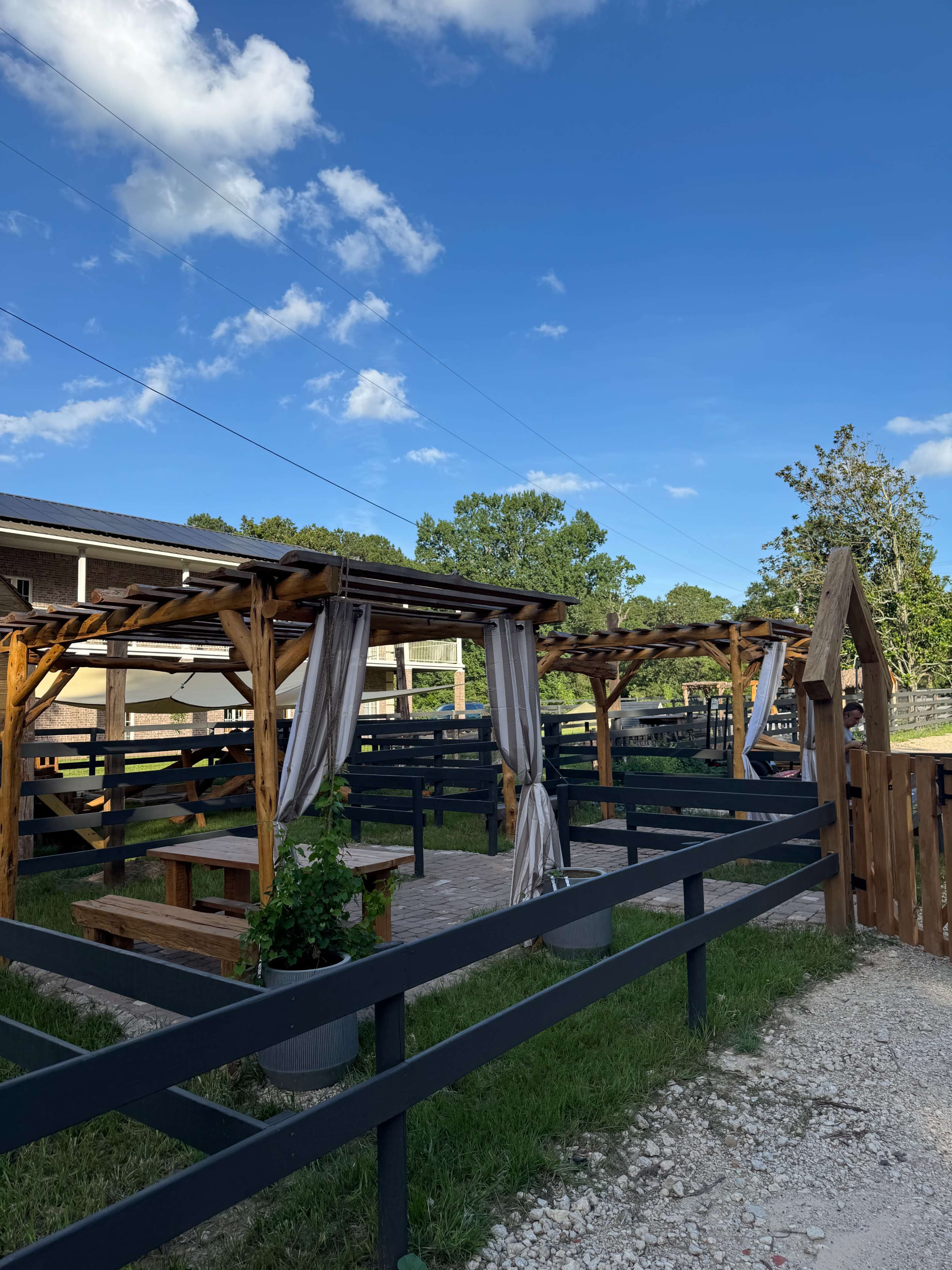 The image shows a wooden outdoor seating area with pergolas and curtains, surrounded by a grass lawn and wooden fencing under a clear blue sky.
