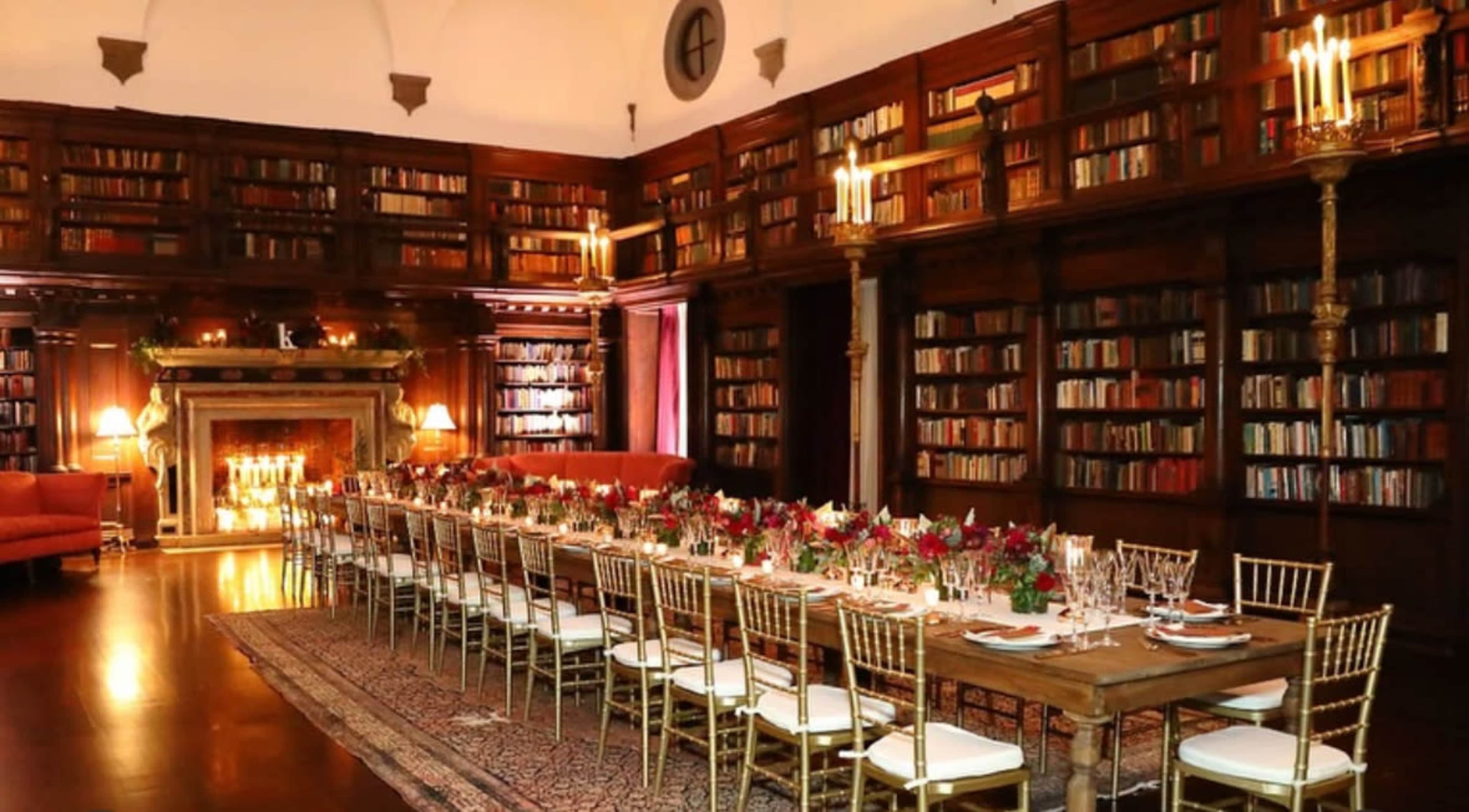 A long dining table is elegantly set in a library filled with bookshelves, with a fireplace and seating along the walls.