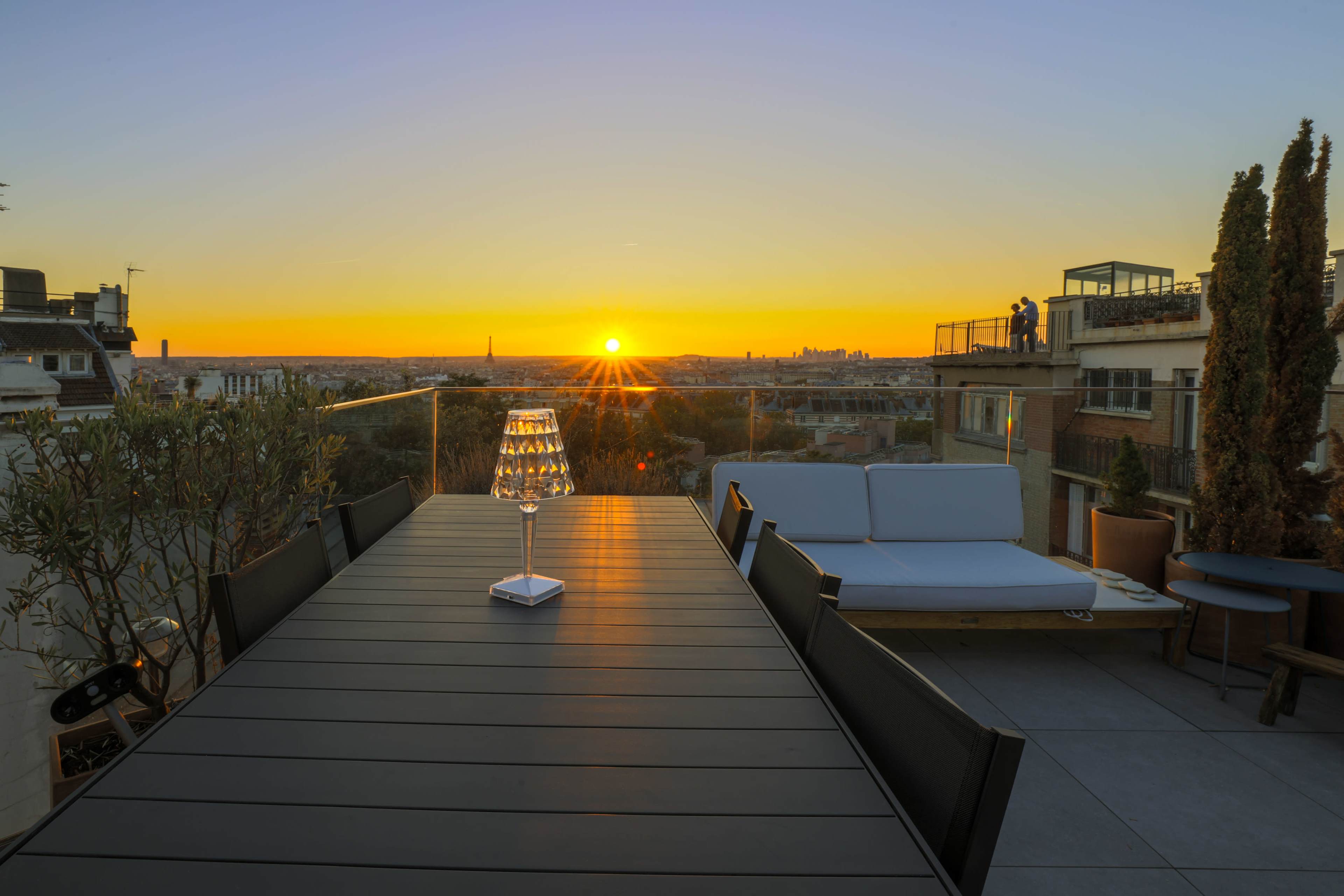 A sleek dining table with a lit candle sits on a rooftop terrace overlooking a city skyline at sunset.