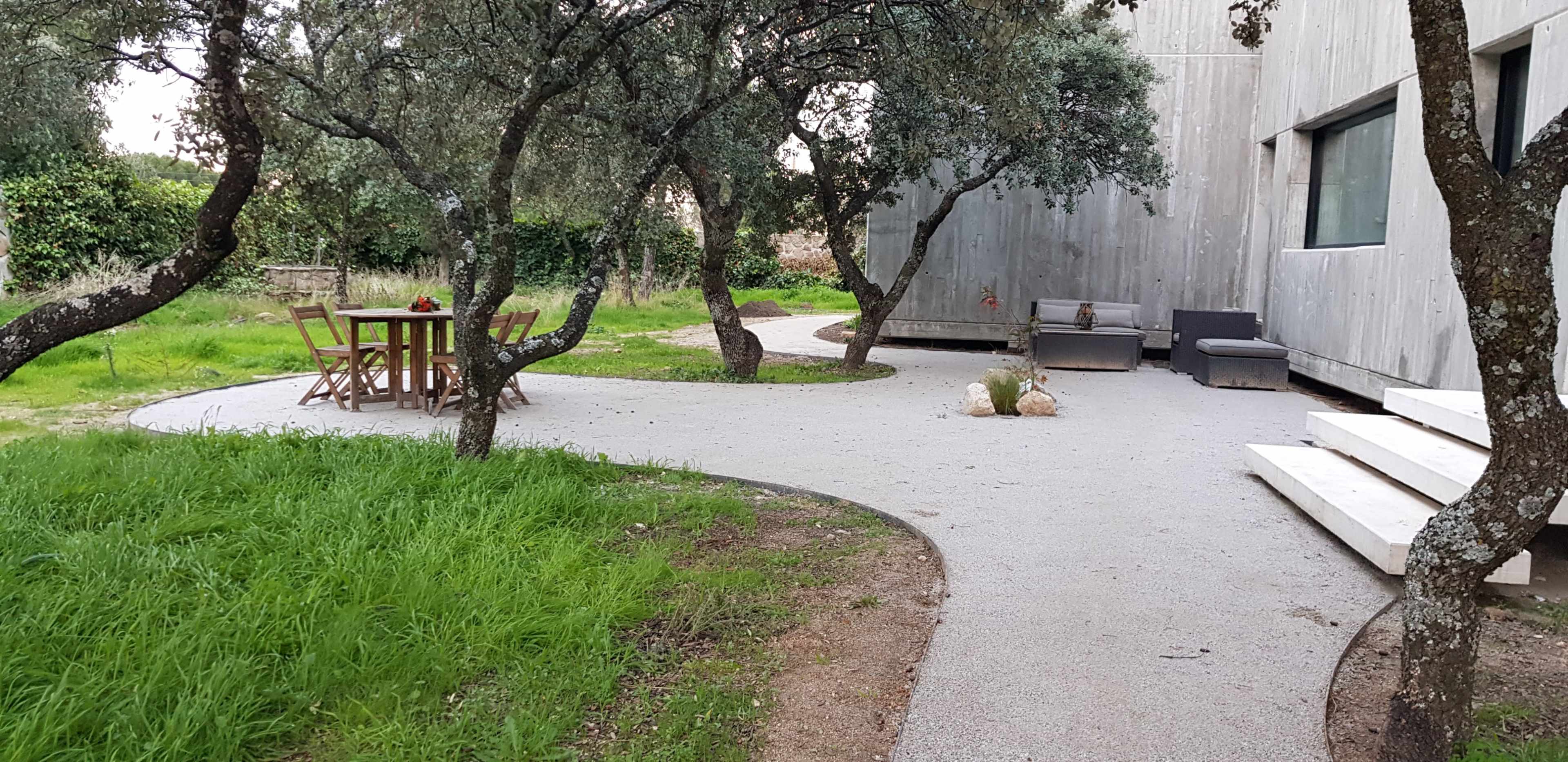 The image shows a modern outdoor space featuring a gravel pathway lined with trees, leading to a wooden dining table set and a seating area near a contemporary building.