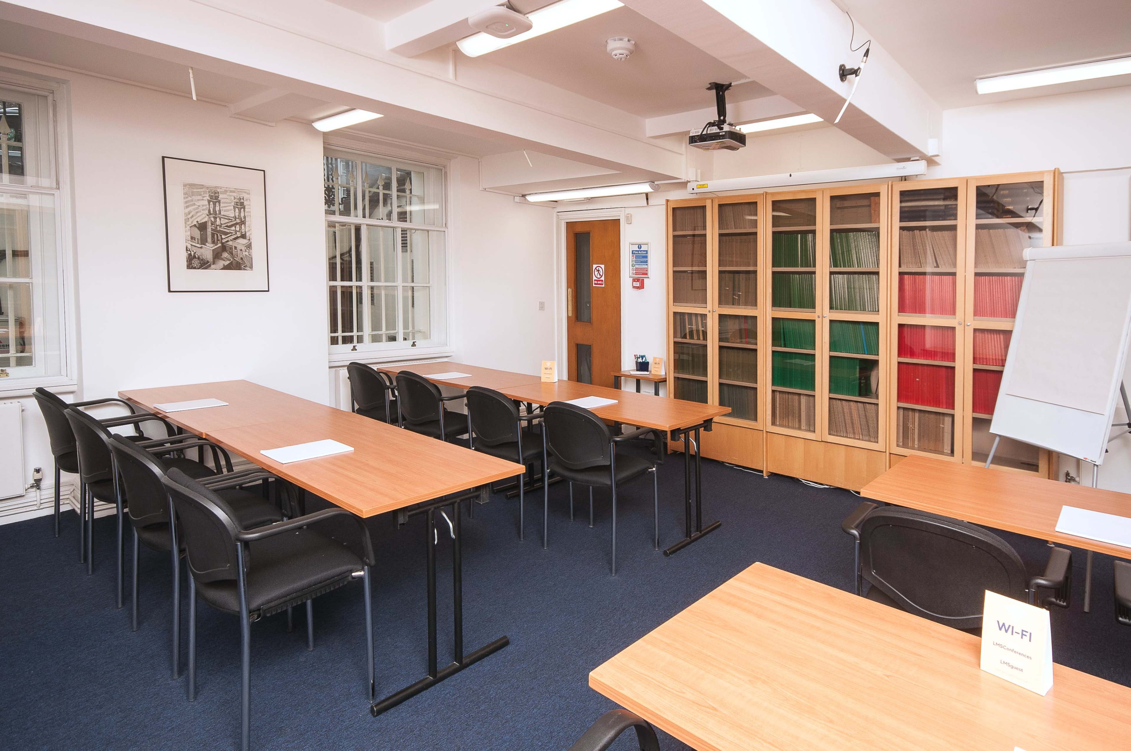 A classroom features rows of wooden tables arranged for an instructional setting, with a large cabinet of colored folders and a projector mounted on the ceiling.