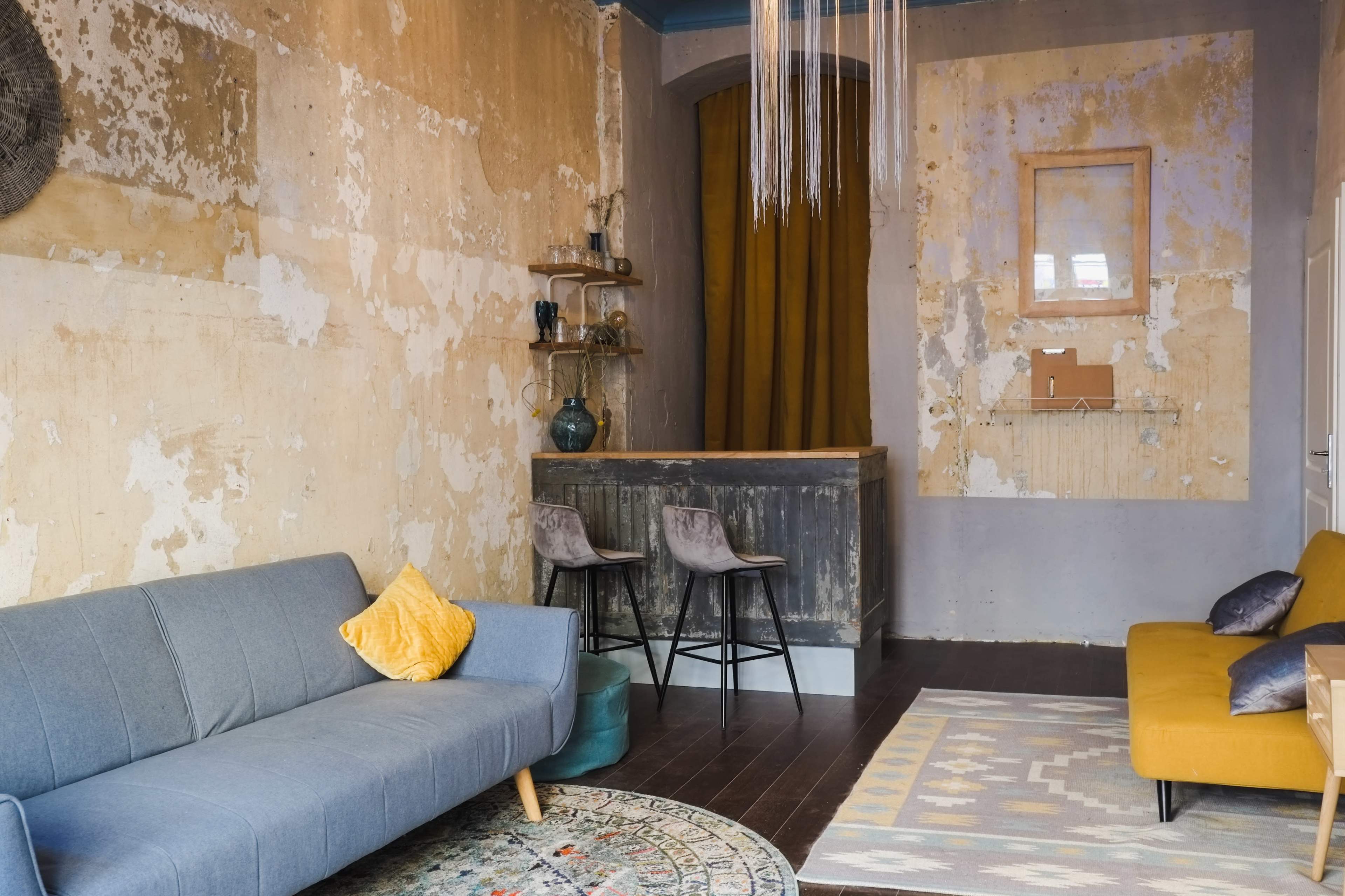 A rustic interior with a gray sofa, an orange couch, a small bar area, and peeling wallpaper.