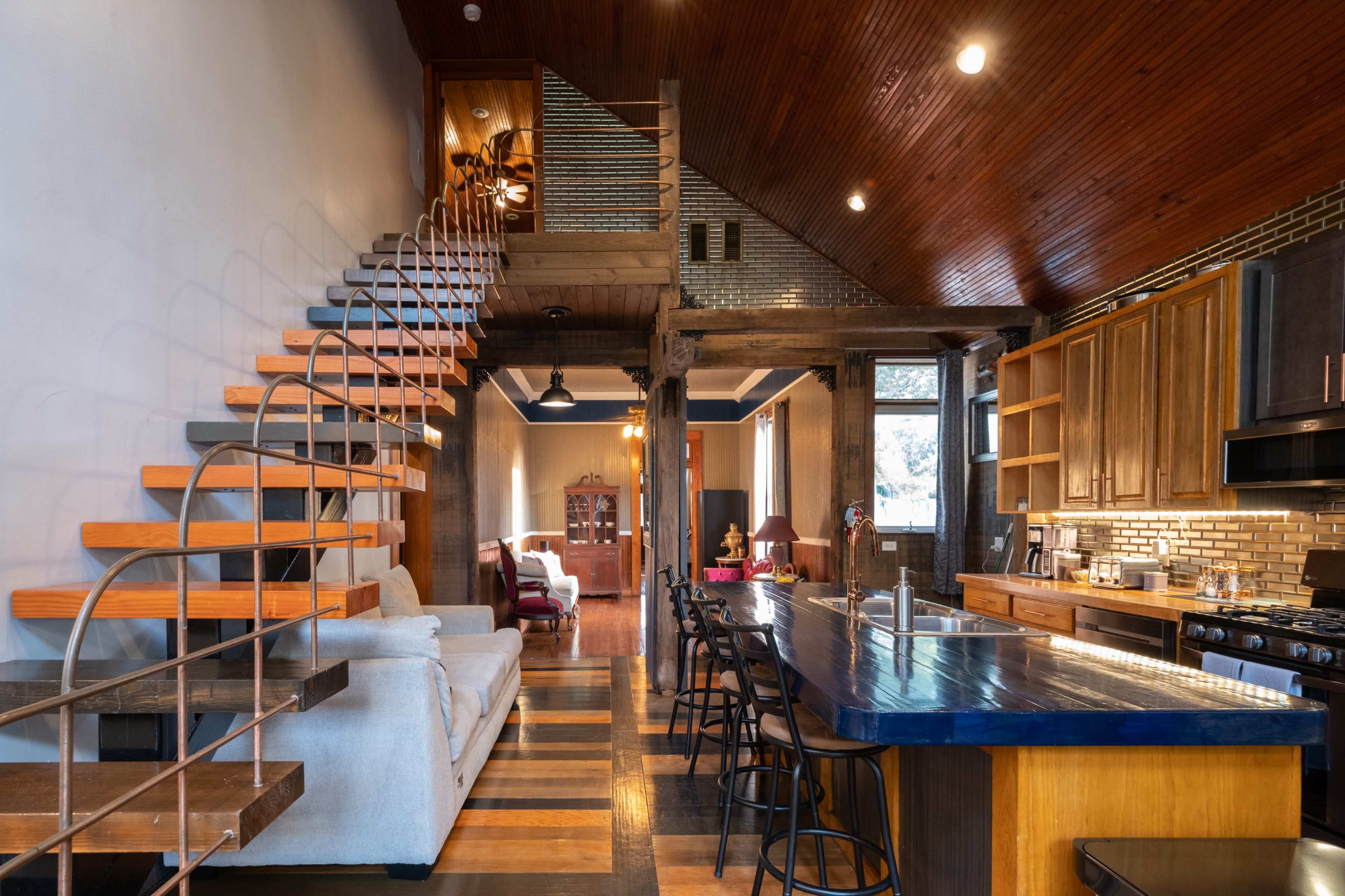 The image shows a modern, open-concept living space with a kitchen island, a staircase leading to a lofted area, and a seating area featuring a sofa.