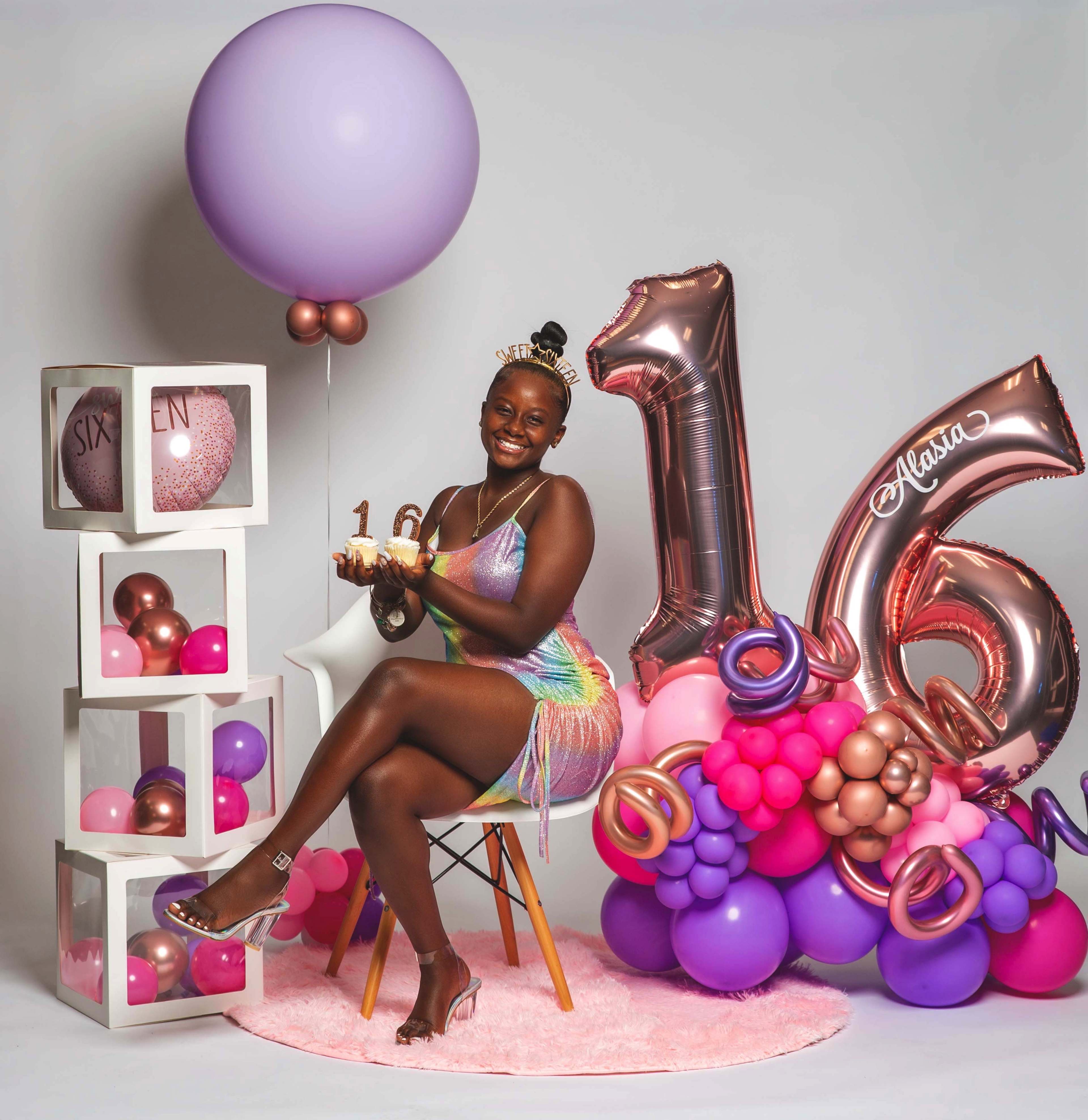 A young person sits on a chair holding a small cake, surrounded by colorful balloons and decorative displays for a sweet sixteen celebration.