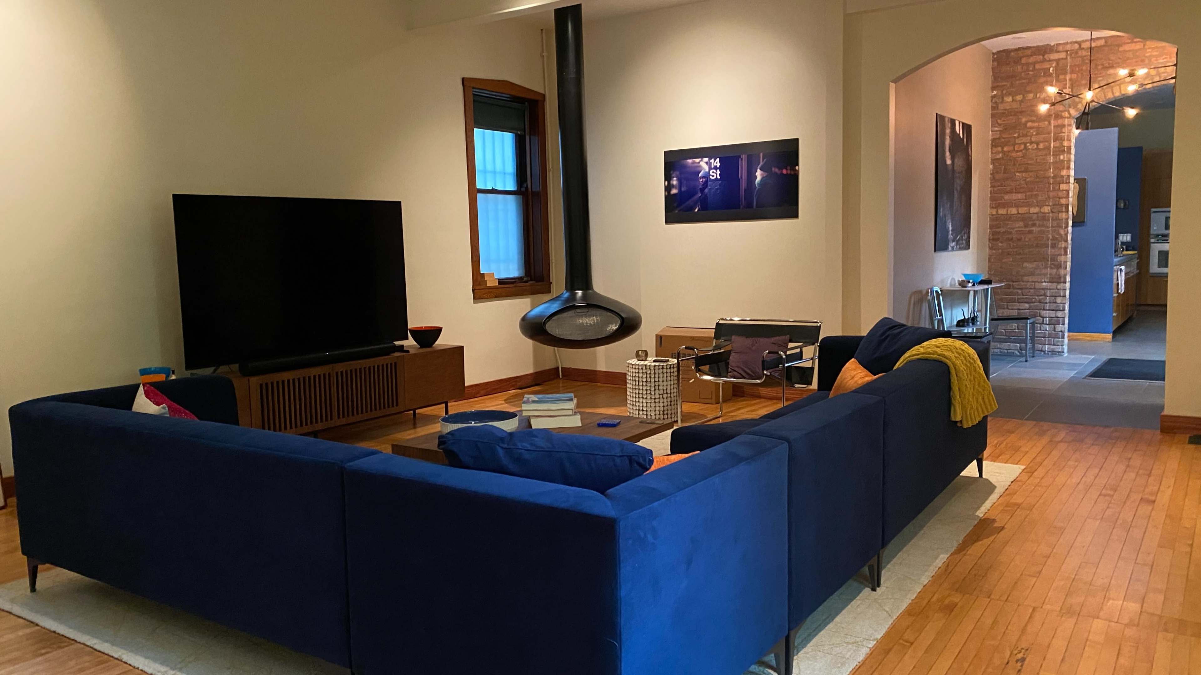 The image shows a modern living room with a blue sectional sofa, a large television on a media console, and an archway leading to a hallway with visible brick walls.