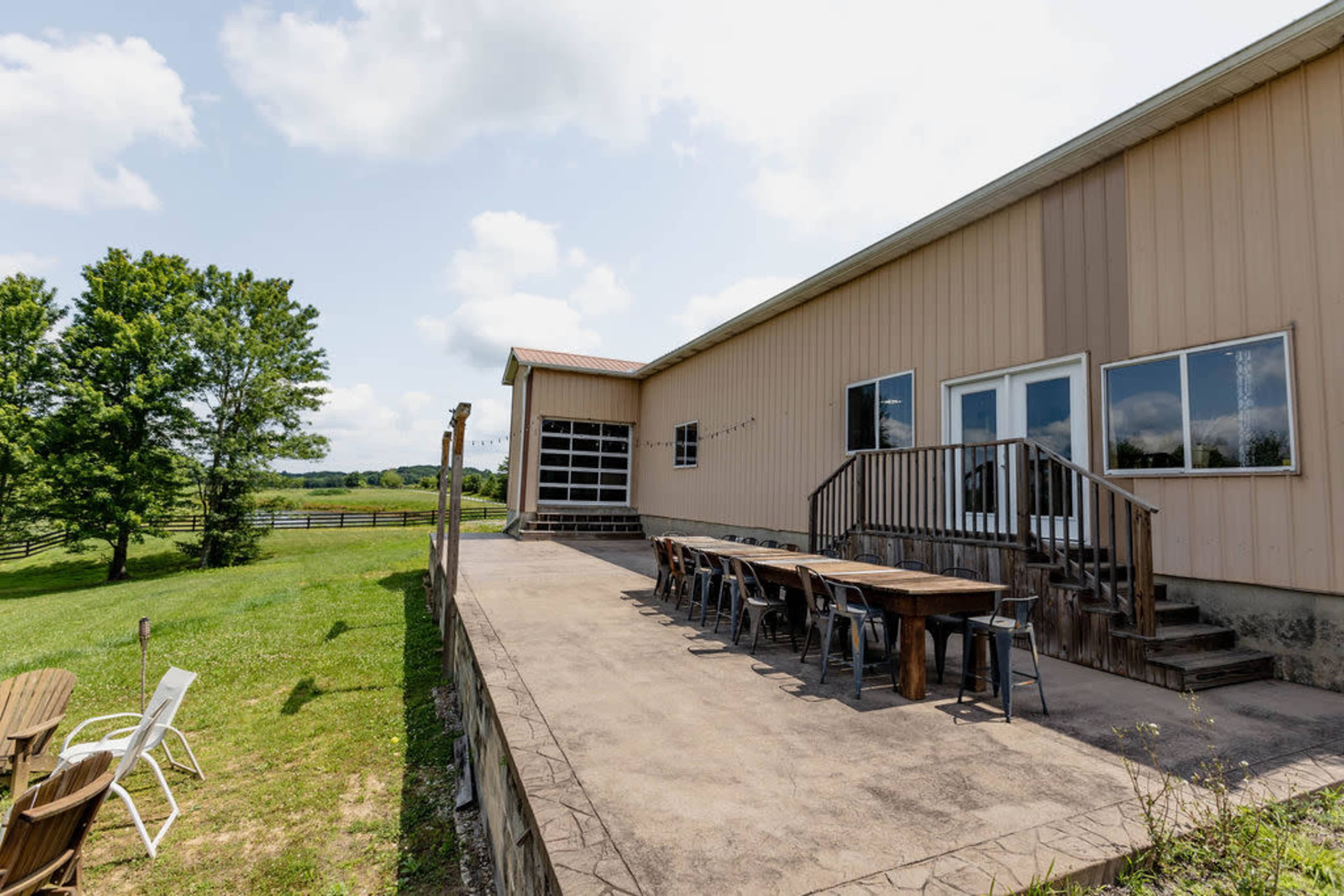 The image shows a large rustic building with a wooden deck and a long table set up outside, surrounded by a grassy area and trees.
