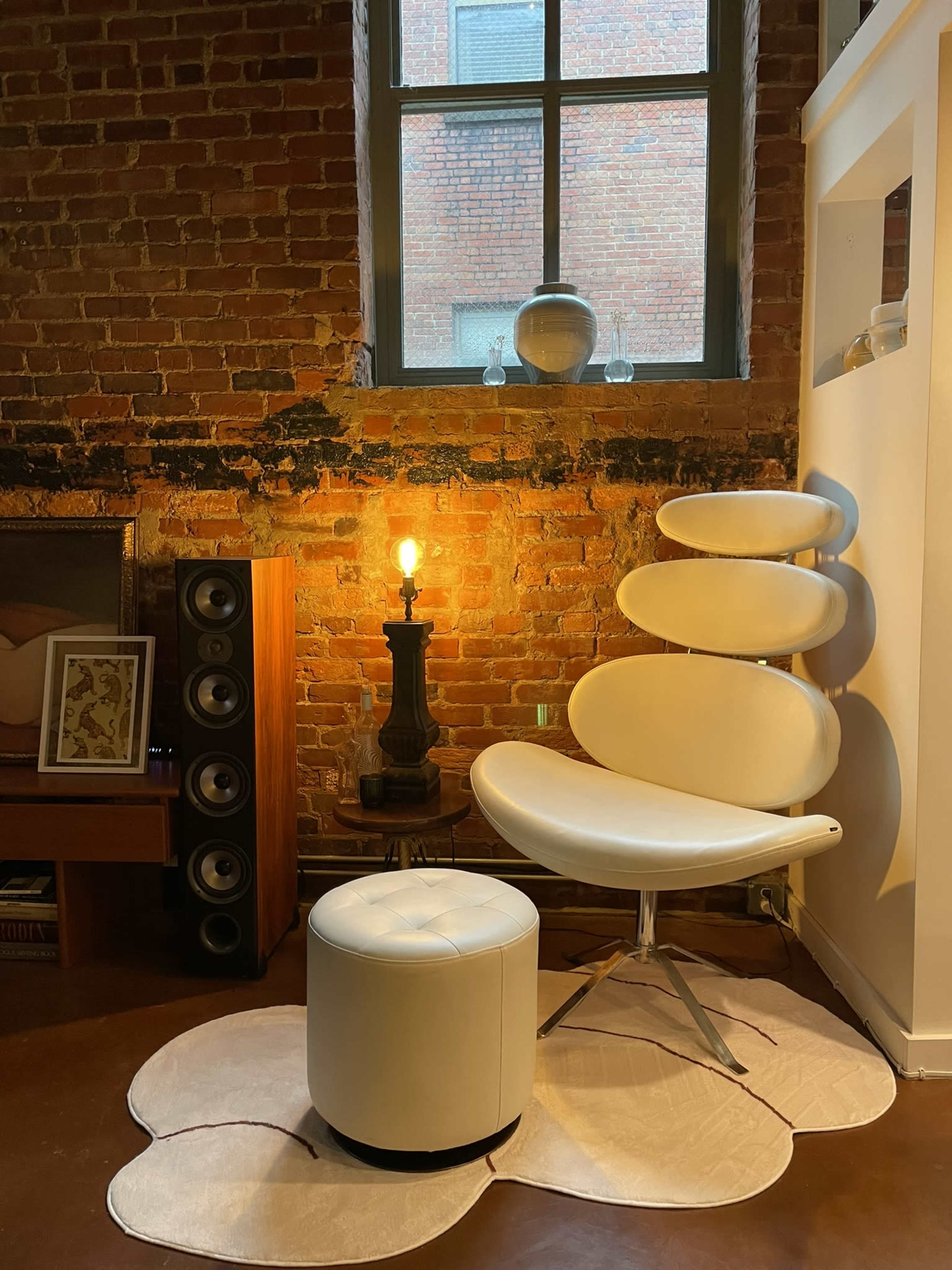 A modern white chair and ottoman are positioned near a window in a brick-walled room, accompanied by a lamp and a speaker setup.