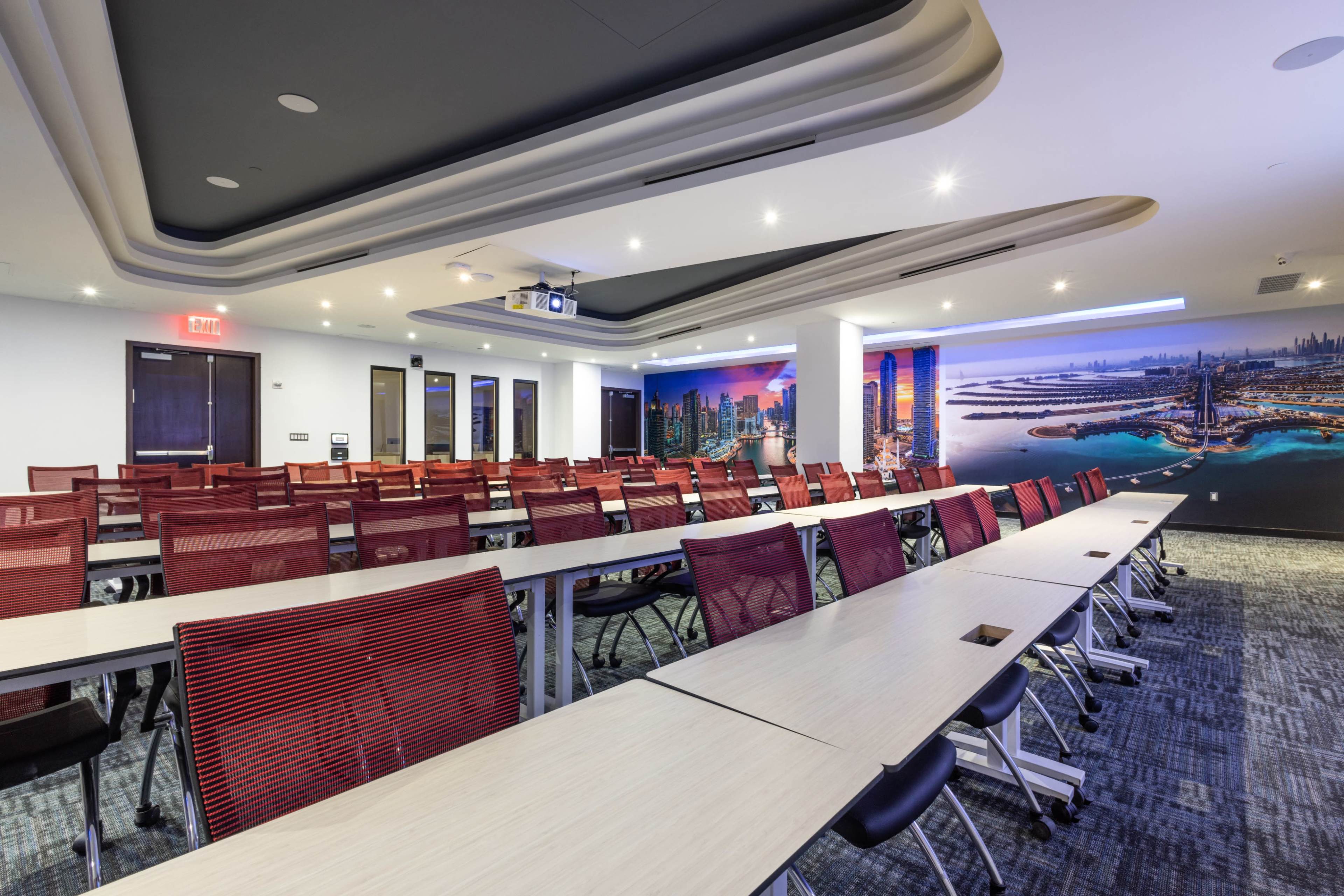 A modern conference room with rows of red chairs and long tables, equipped with a projector and featuring a large mural of a city skyline on one wall.