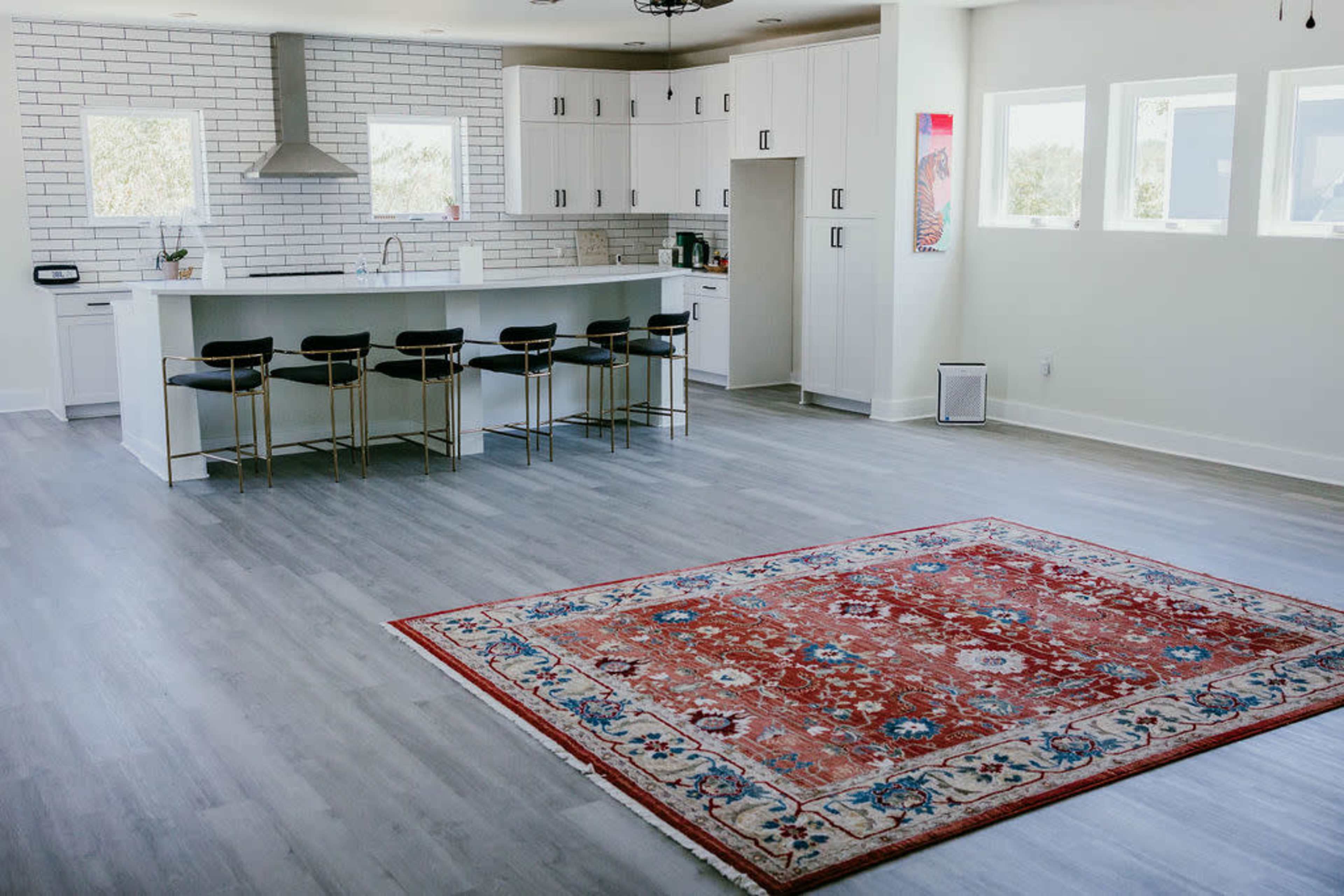 The image shows a bright, minimalist kitchen and living area featuring a large area rug on a light gray flooring, with a kitchen island and bar stools against a wall of cabinets.