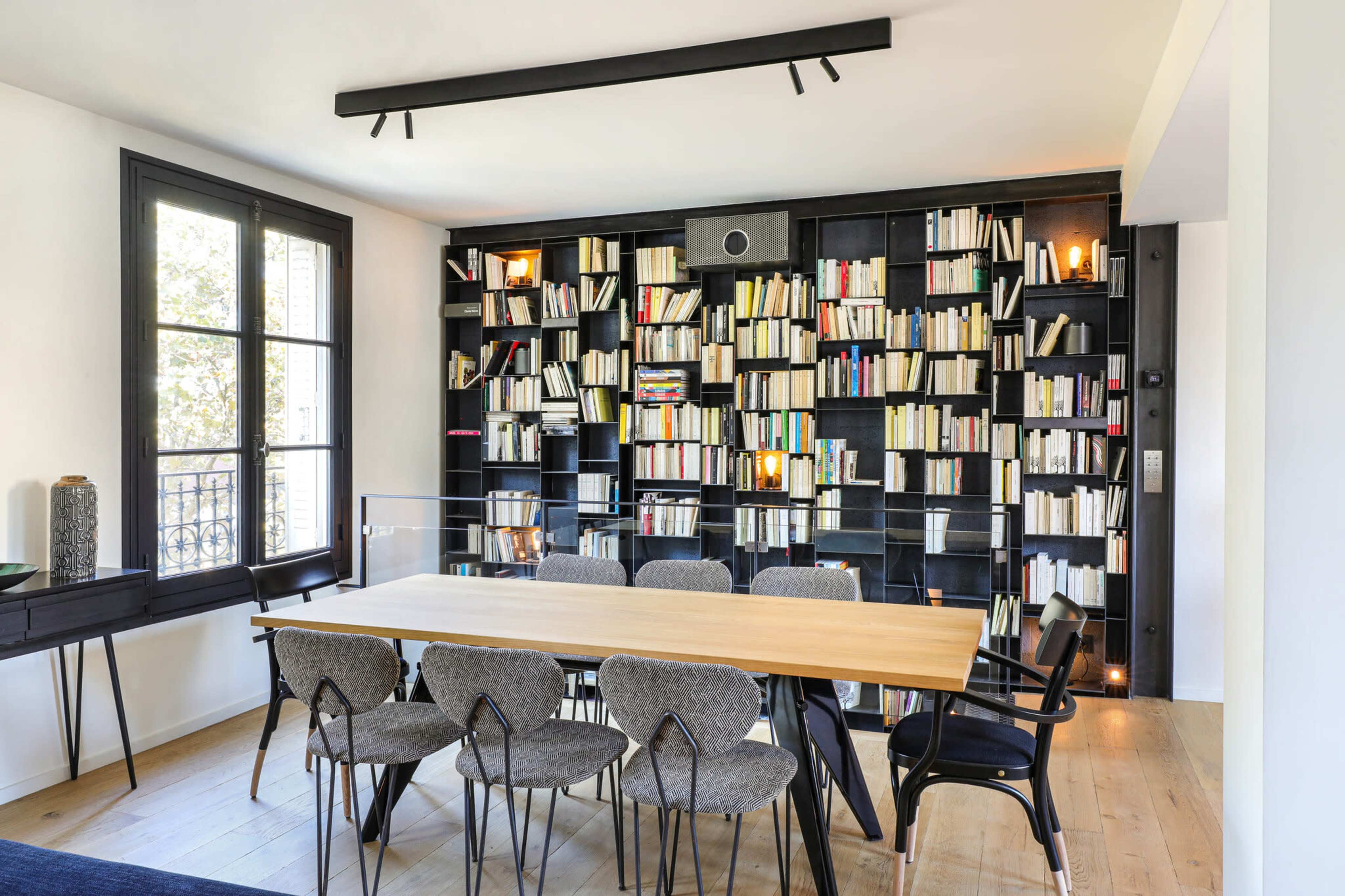 A modern dining area features a long wooden table surrounded by upholstered chairs, with a large bookshelf filled with white and black books on one wall.