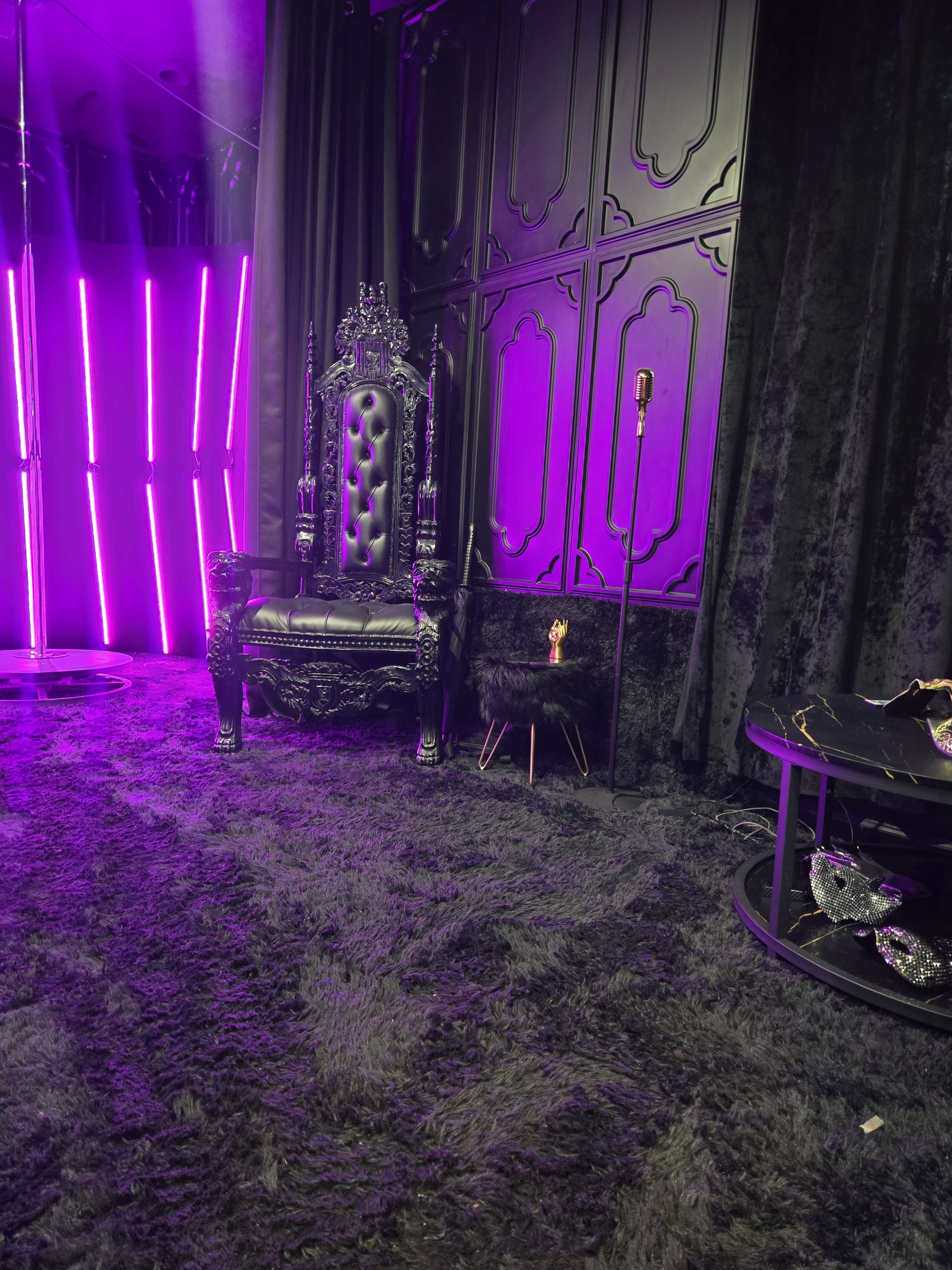 The image shows a dark room featuring a large, ornate black throne, a modern standing lamp, and a round table with decorative items, all set against a textured black background and illuminated by purple lighting.