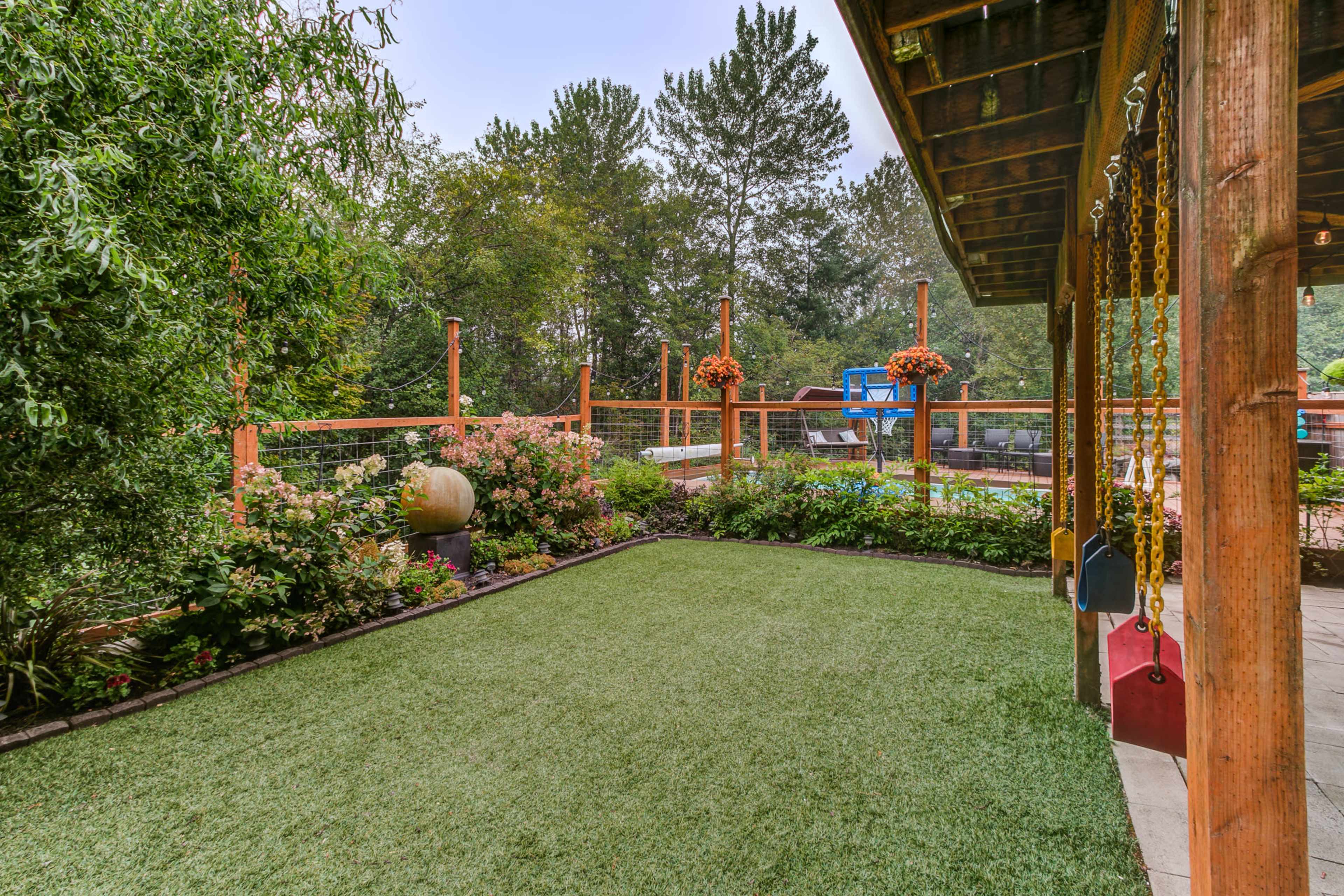 The image shows a landscaped backyard with synthetic grass, colorful flower beds, a wooden deck, and a swing set.