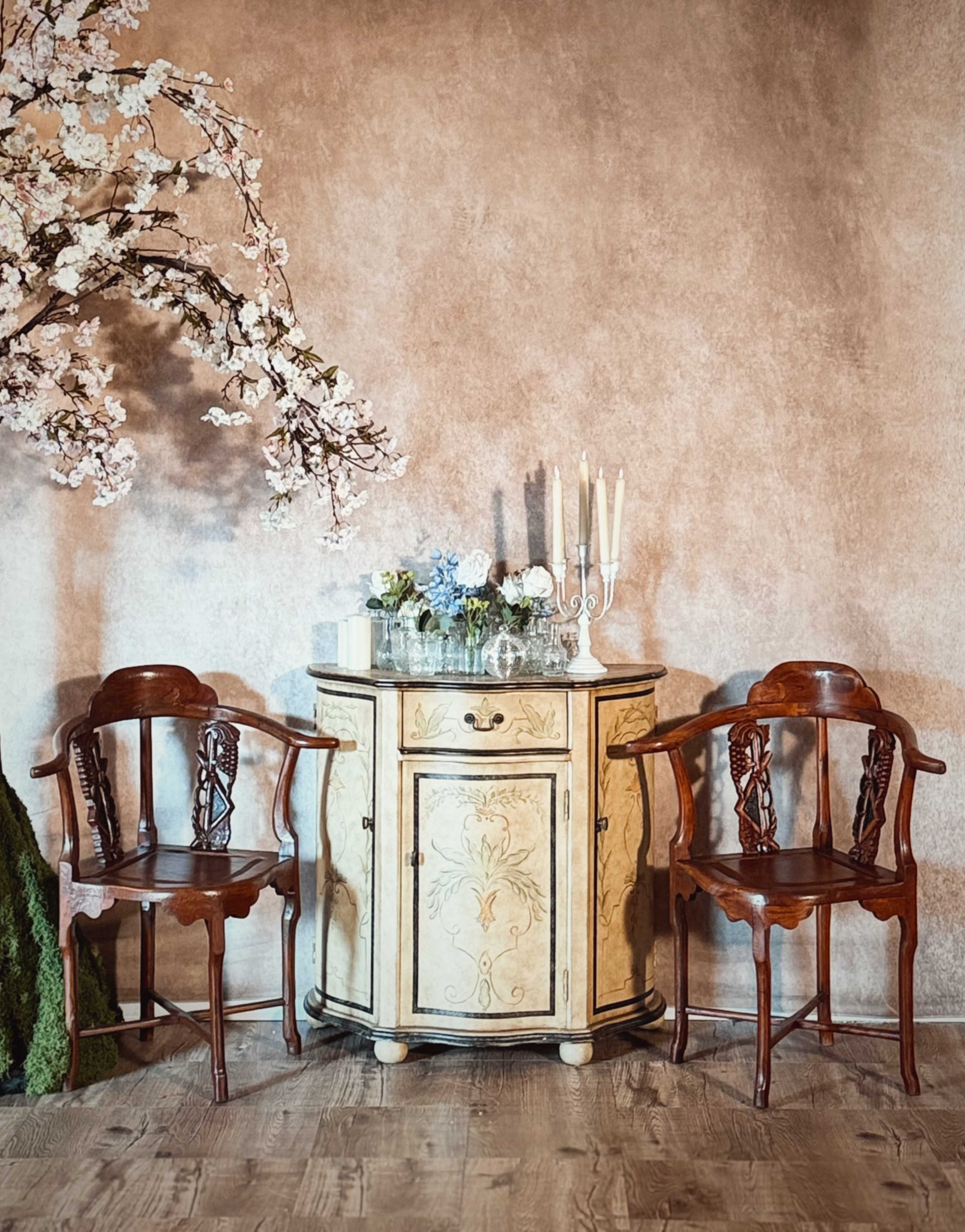 The image shows a small round table with decorative carvings and a glass top, flanked by two wooden chairs and a cherry blossom tree in a softly lit room.