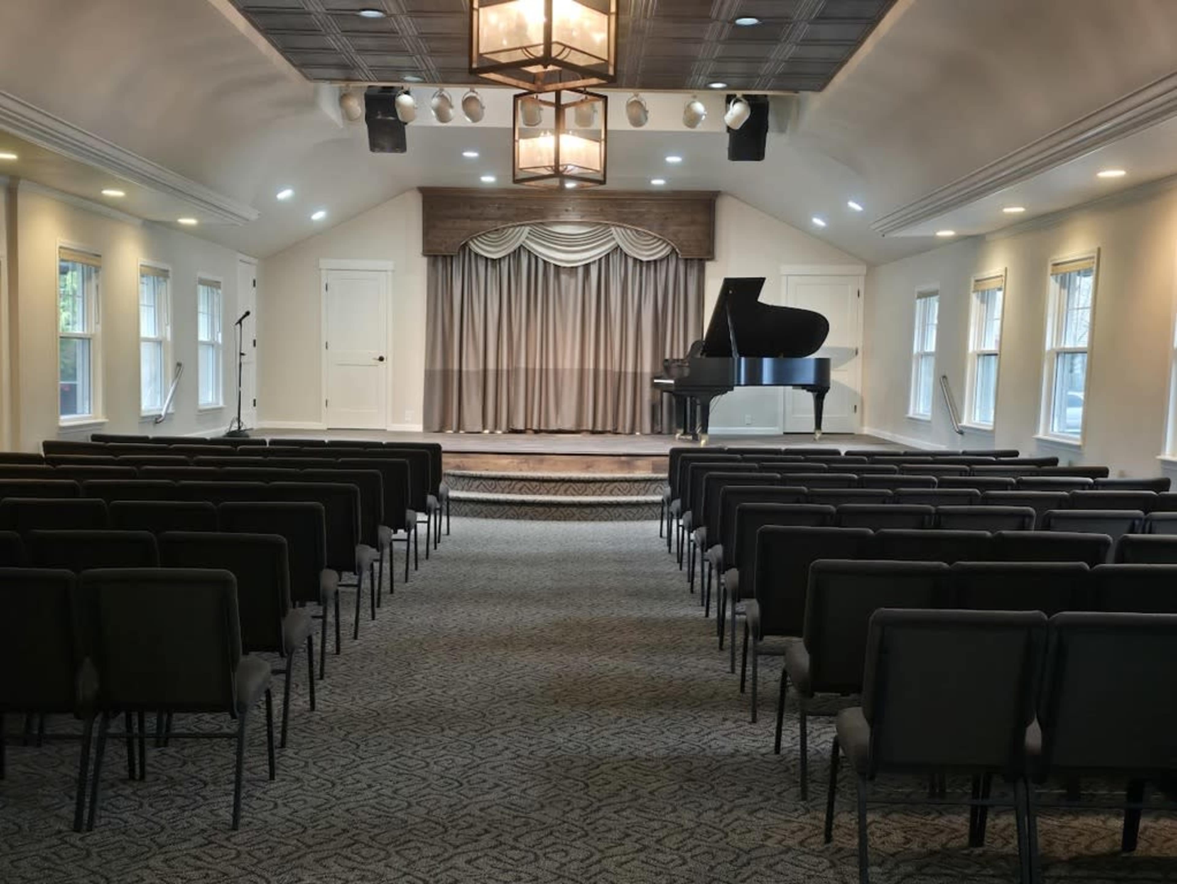The image shows an indoor performance space with rows of black chairs facing a stage that features a piano and a large curtain backdrop.
