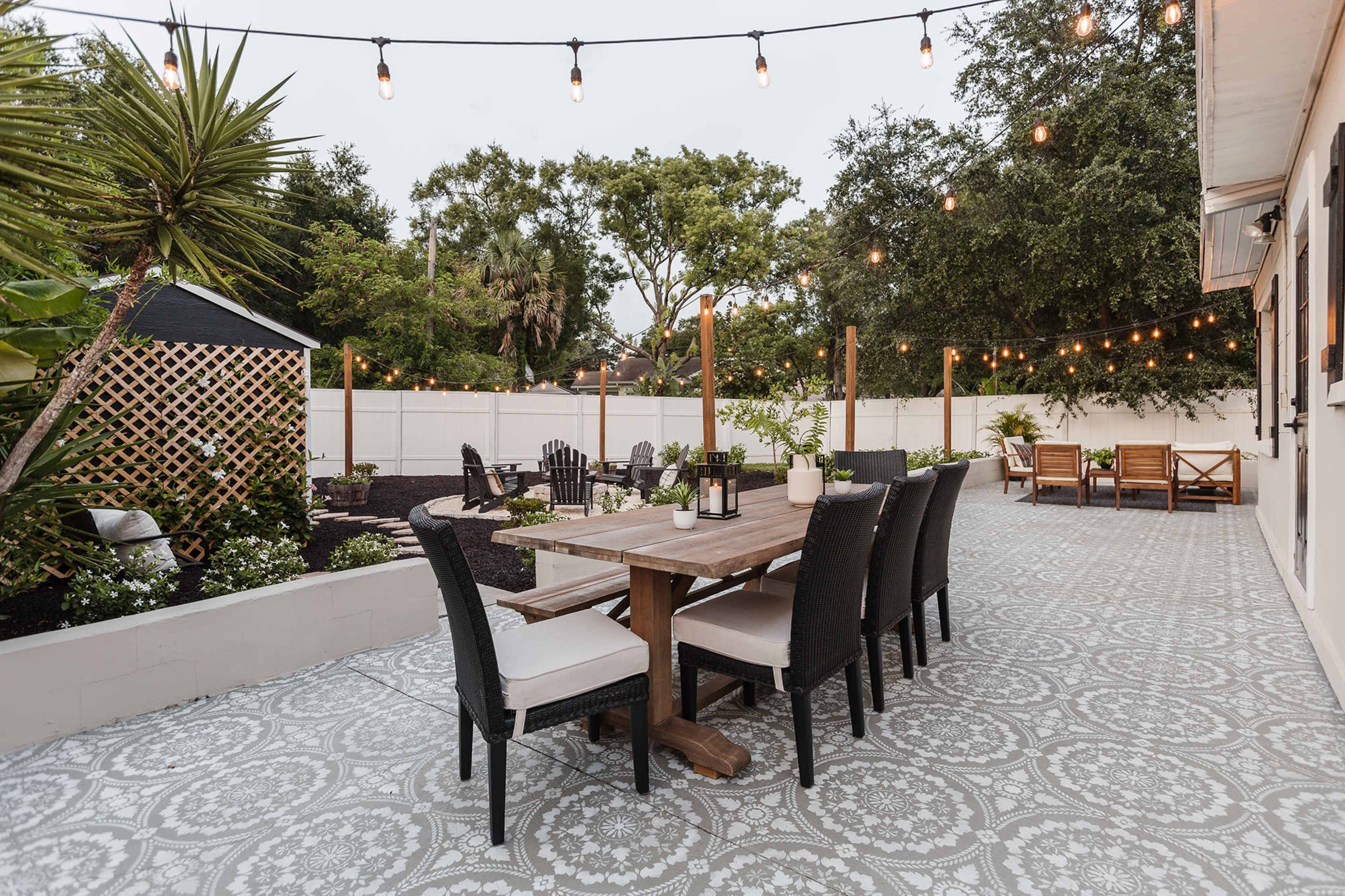 A spacious outdoor patio features a large wooden dining table surrounded by black chairs, with hanging string lights and greenery in the background.