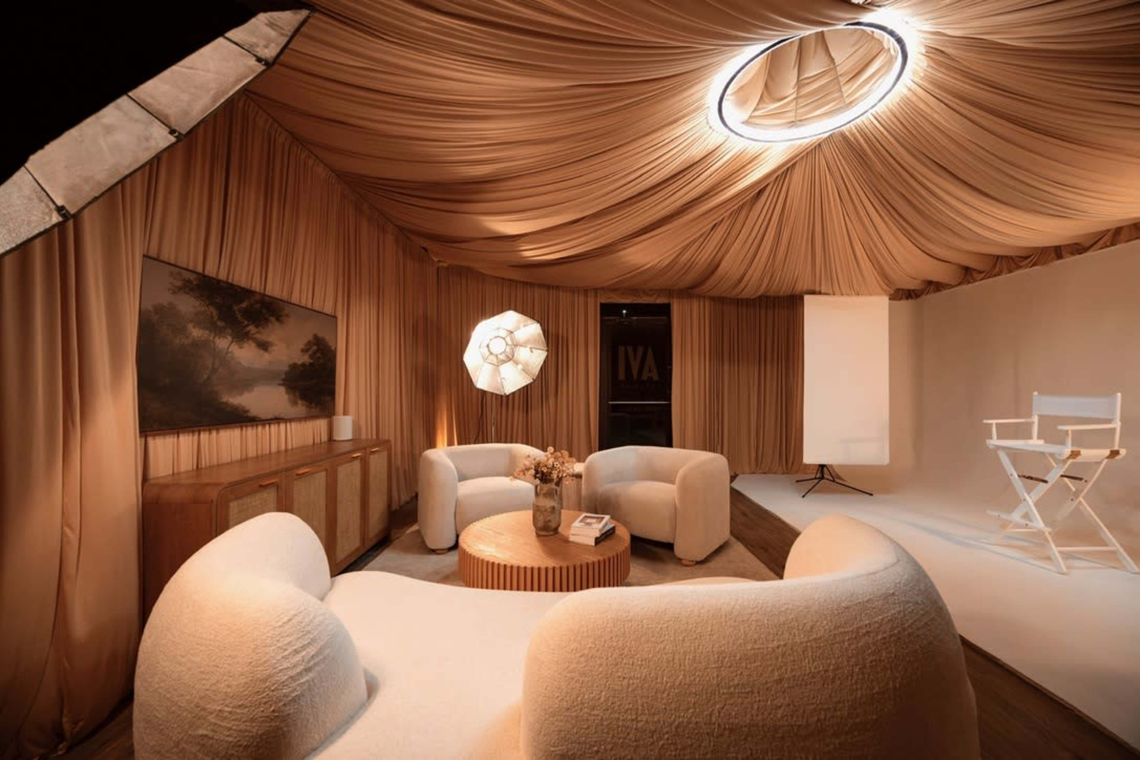 The image shows a softly-lit room with a draped ceiling, featuring a circular seating arrangement, a round table, and a backdrop for presentations.