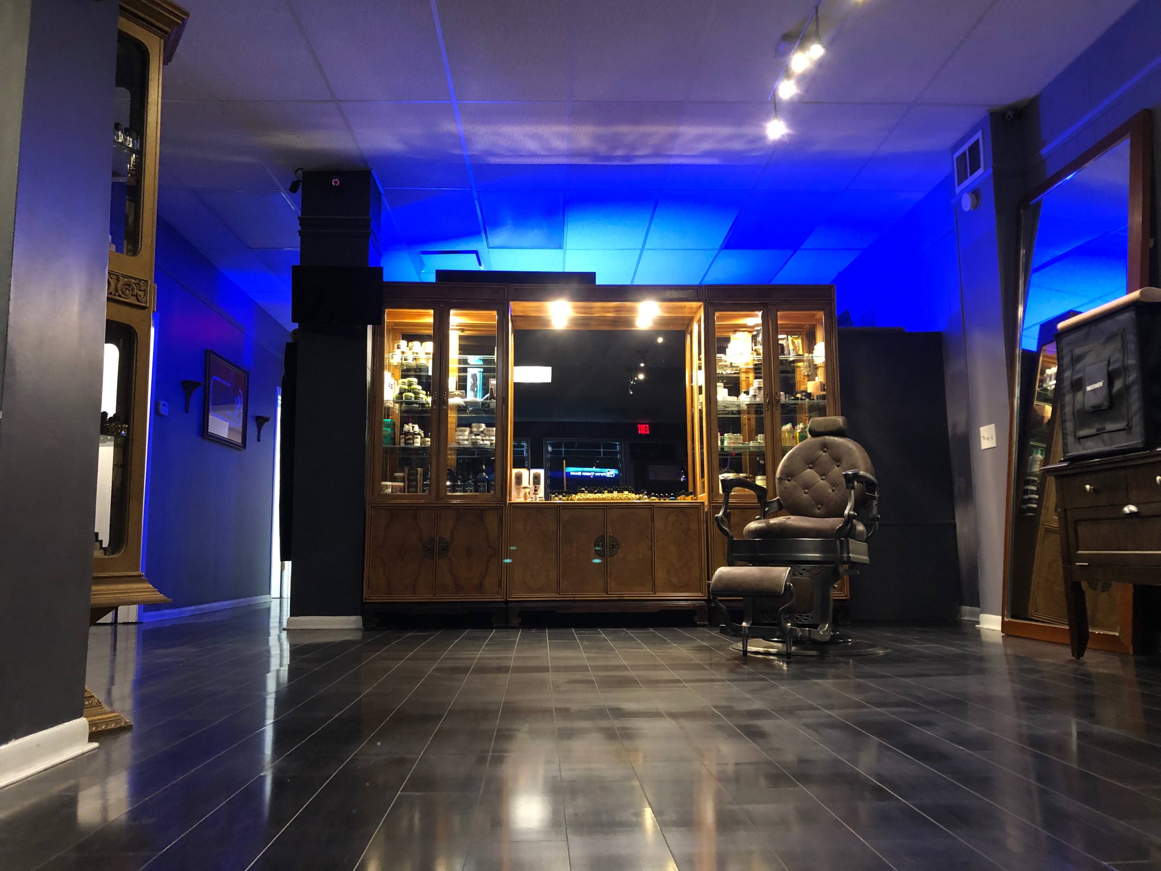 A modern barber shop interior featuring a vintage barber chair, display cabinets filled with products, and blue ambient lighting.
