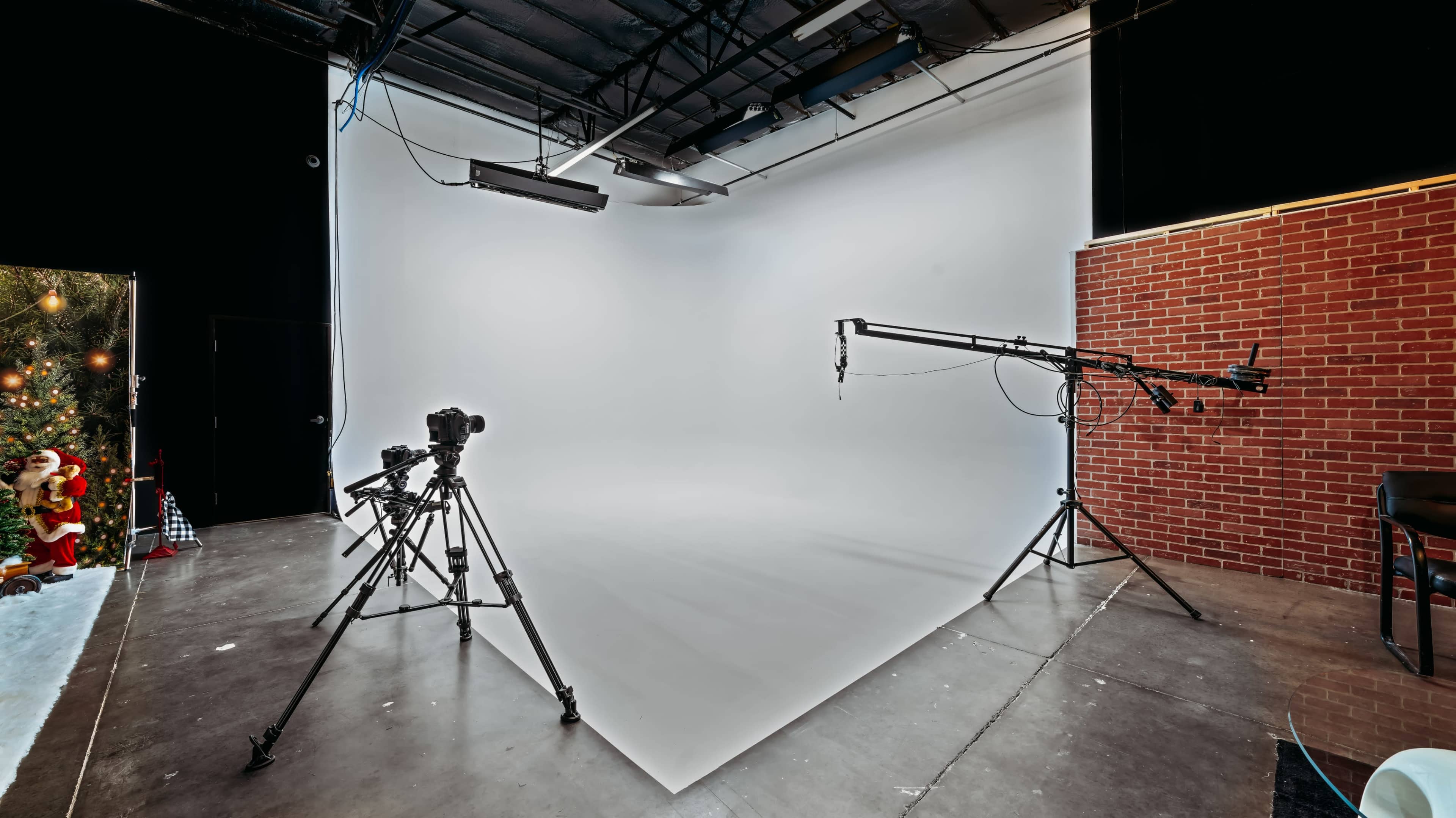 A photography studio with a clean white backdrop, two tripods with cameras, and a ceiling-mounted camera crane, next to a brick wall.