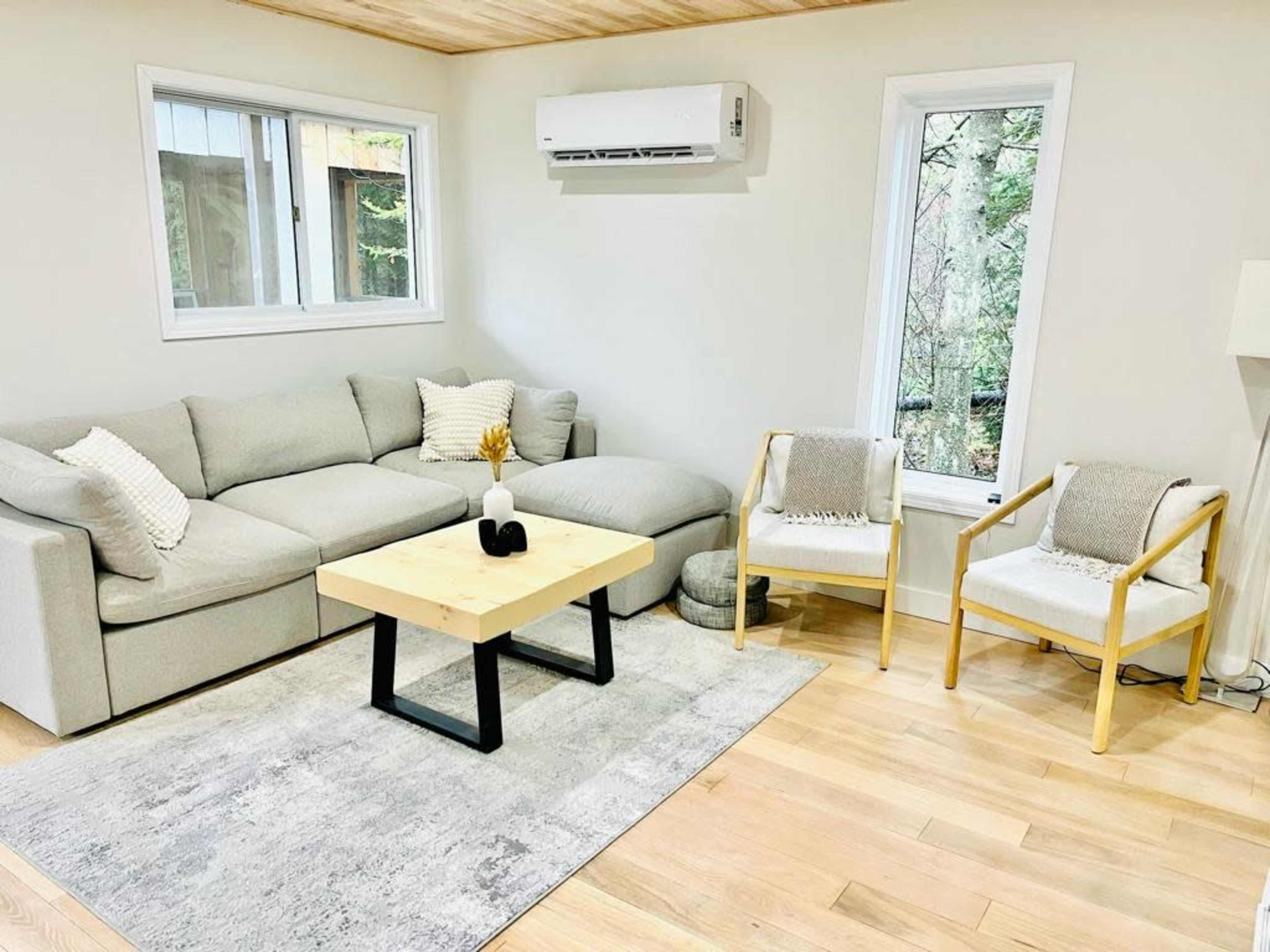The image shows a cozy living room featuring a light gray sectional sofa, two chairs, a wooden coffee table, and large windows allowing natural light.