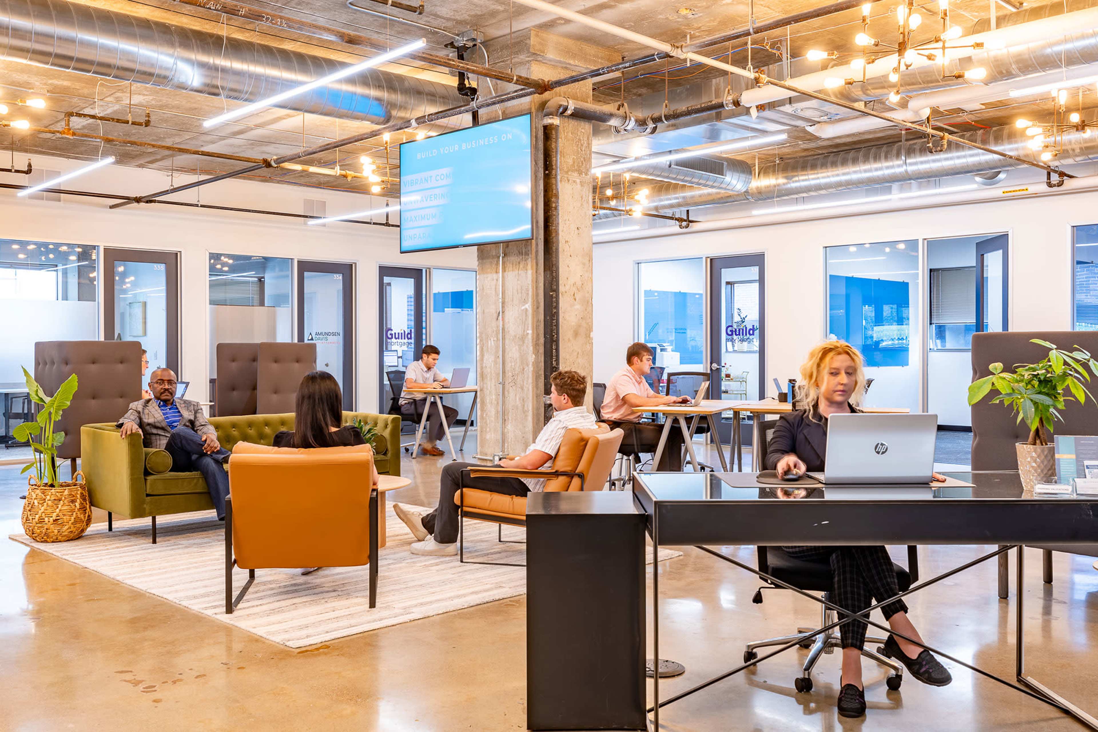 The image shows a modern office space with several people working at desks, lounging on chairs, and collaborating in a well-lit environment featuring large windows and contemporary furniture.