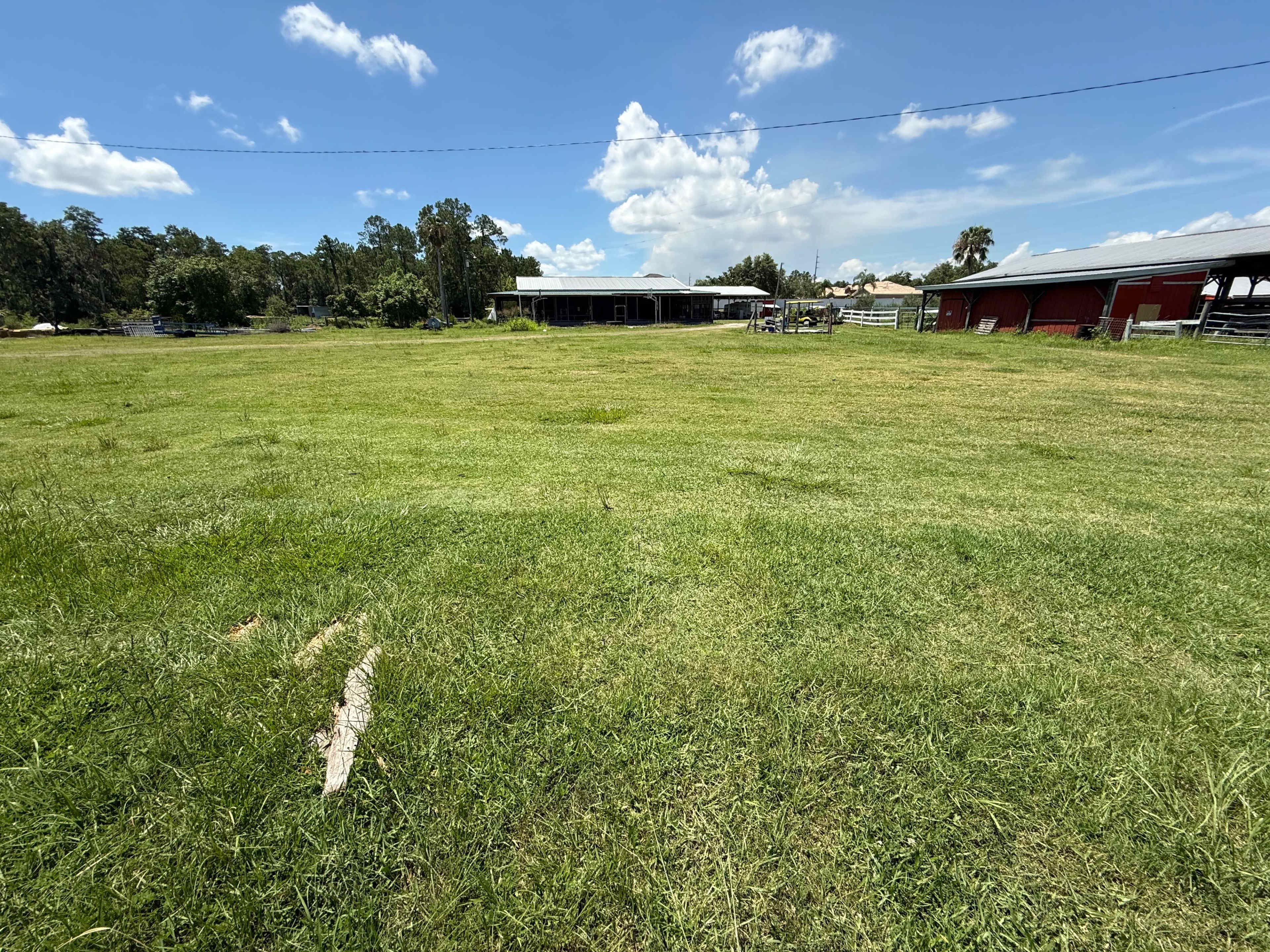The image shows a wide expanse of green grass in a field with several structures, including barns, in the background under a partly cloudy sky.