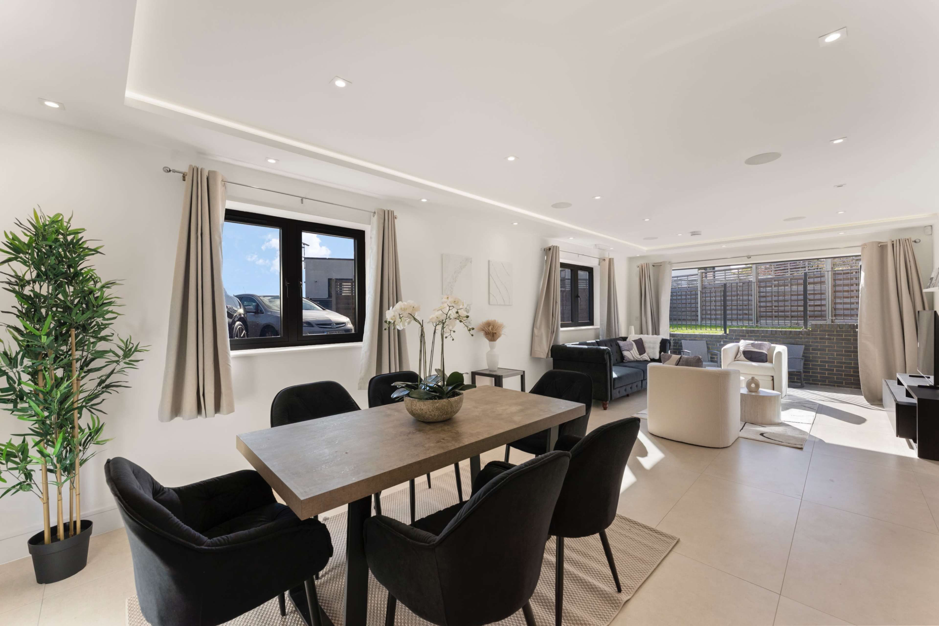 A modern open-concept living and dining area features a wooden table surrounded by black chairs, with large windows letting in natural light and a view of a car parked outside.