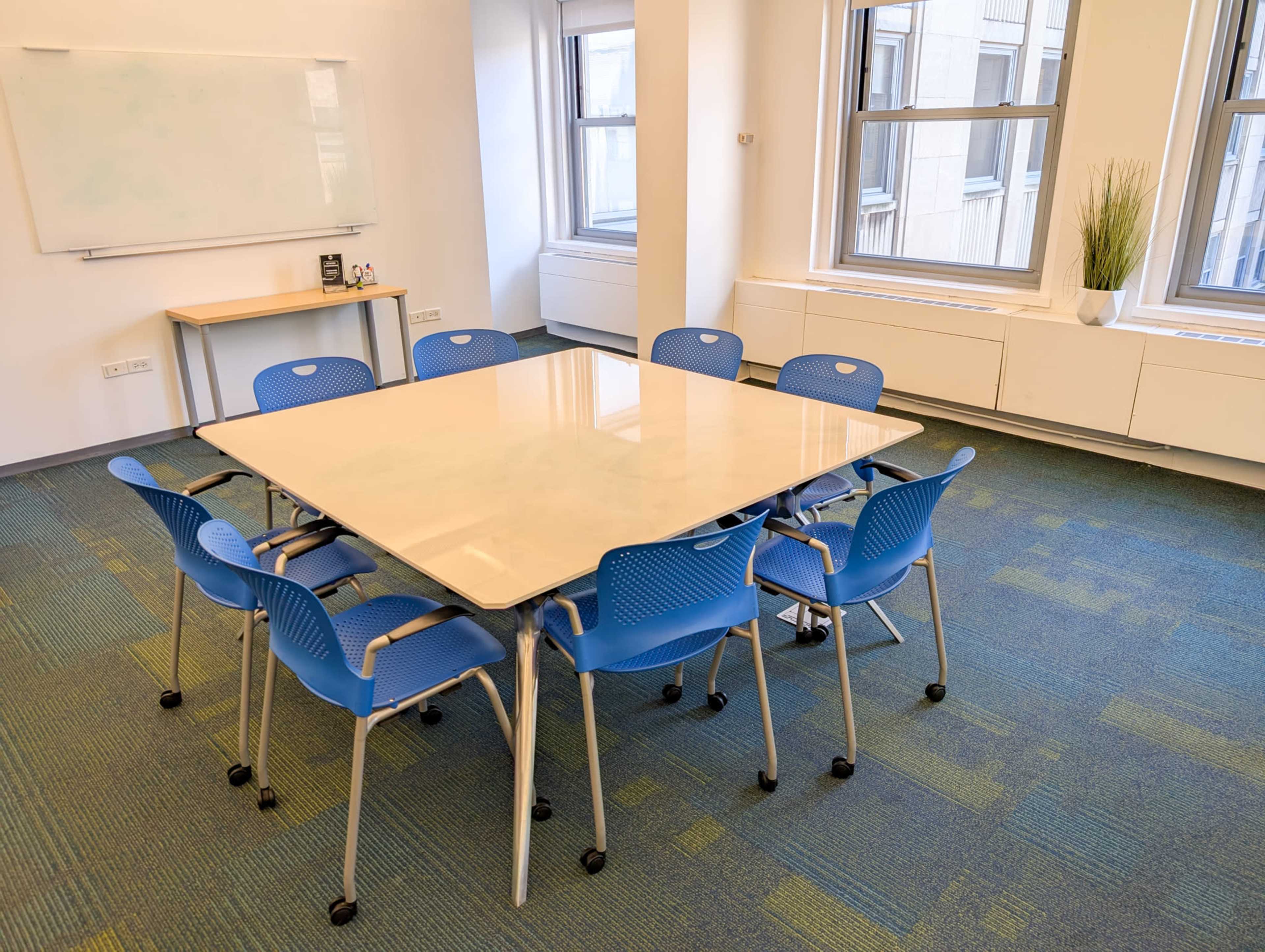 A conference room features a large rectangular table surrounded by eight blue chairs, with a whiteboard and windows in the background.