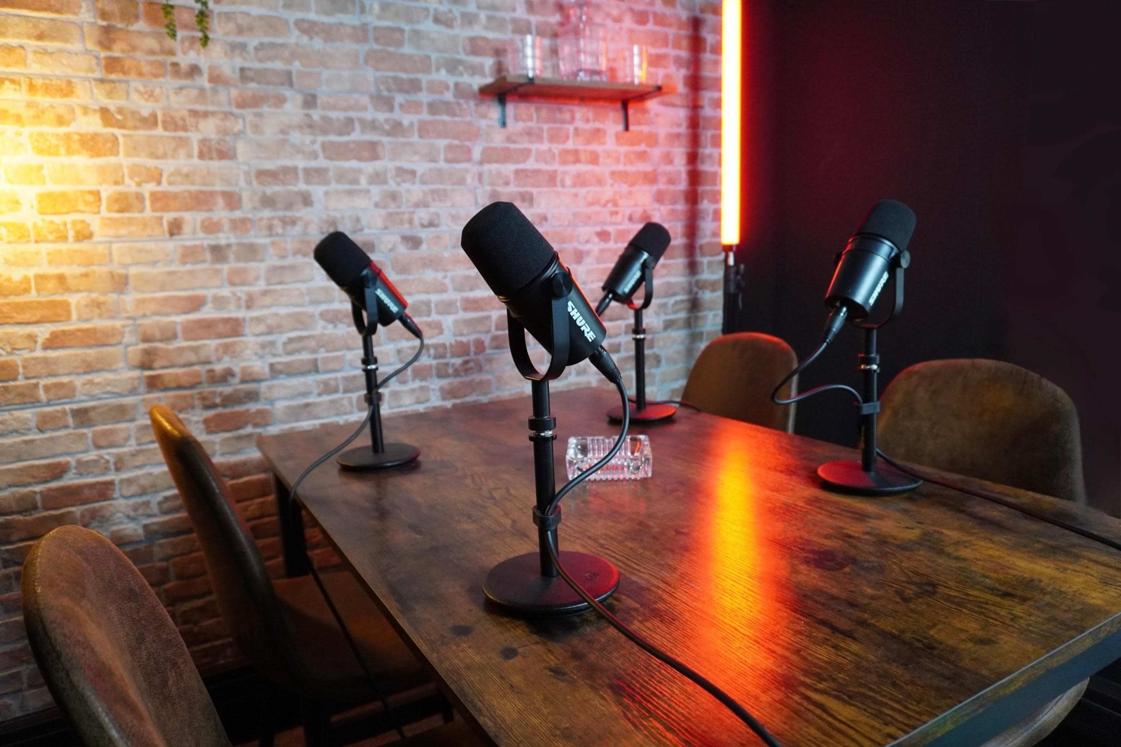 A wooden table with four microphones on stands is arranged in front of a brick wall, illuminated by a neon light.