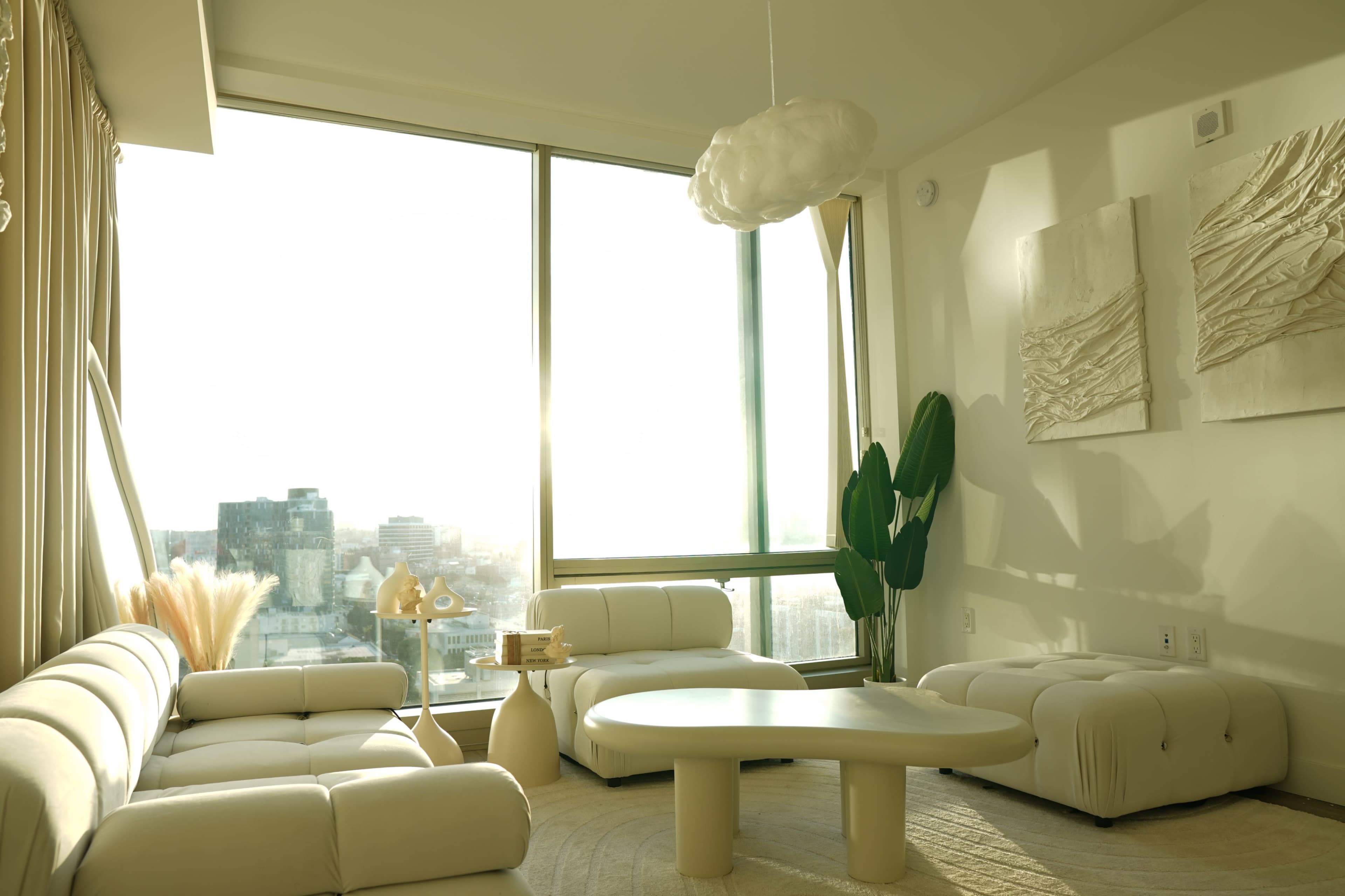 The image shows a modern living room with white furniture, large windows allowing sunlight to stream in, and a decorative potted plant.