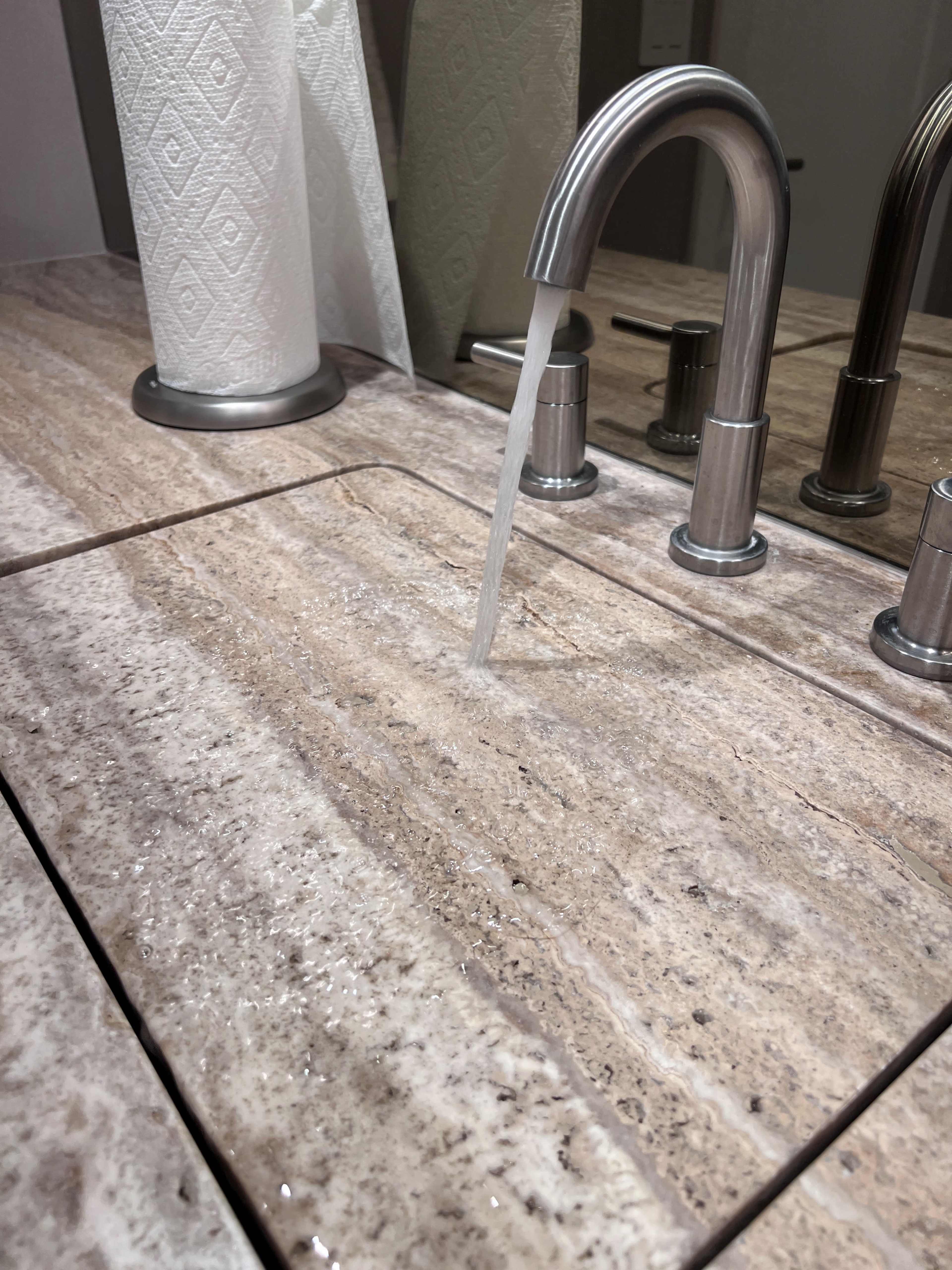 A faucet is releasing a steady stream of water onto a patterned countertop with a paper towel roll in the background.