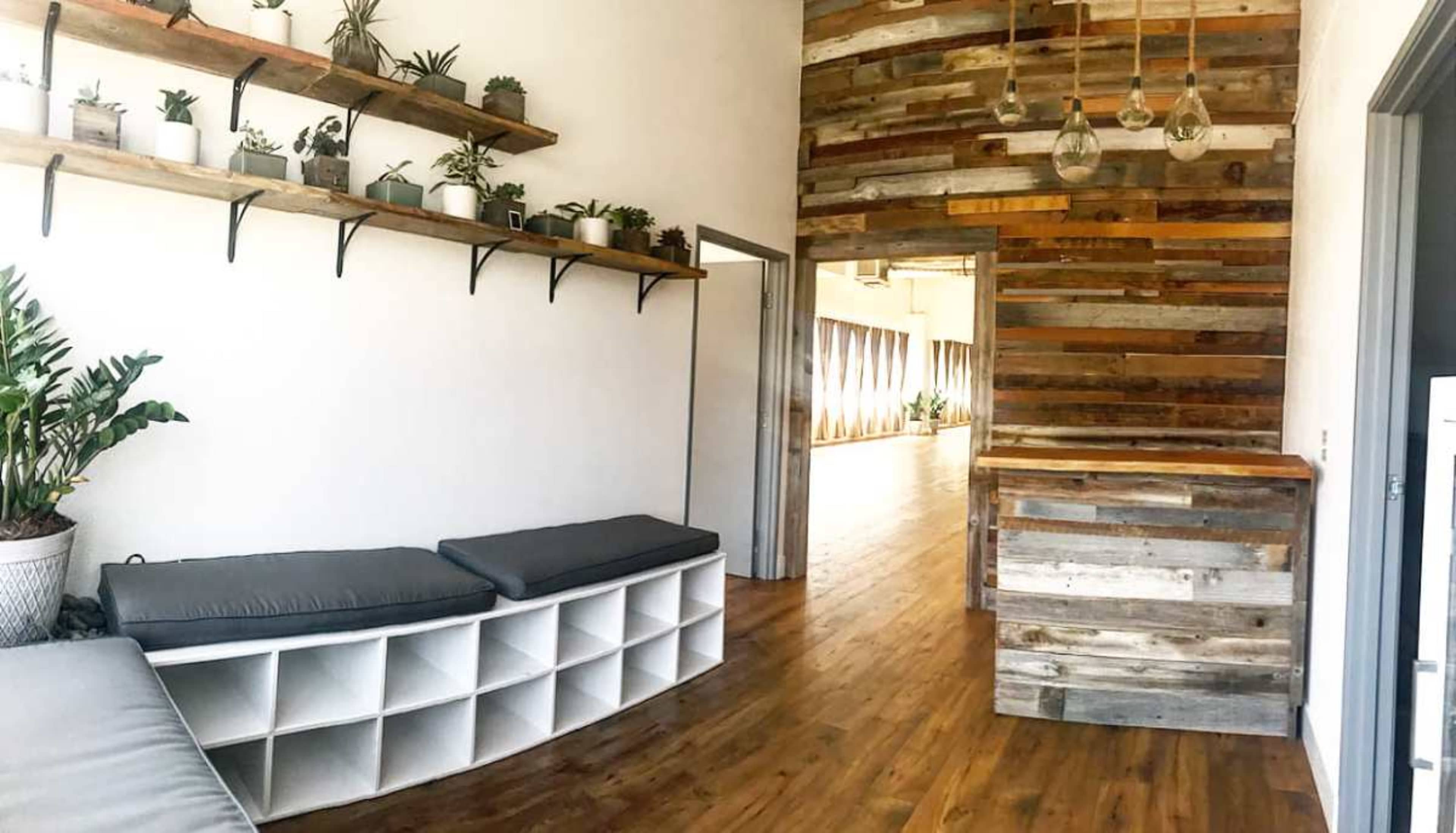 The image shows a modern, minimalistic room with wooden flooring, a bench with cushions, and a wall made of reclaimed wood, along with shelves displaying potted plants.