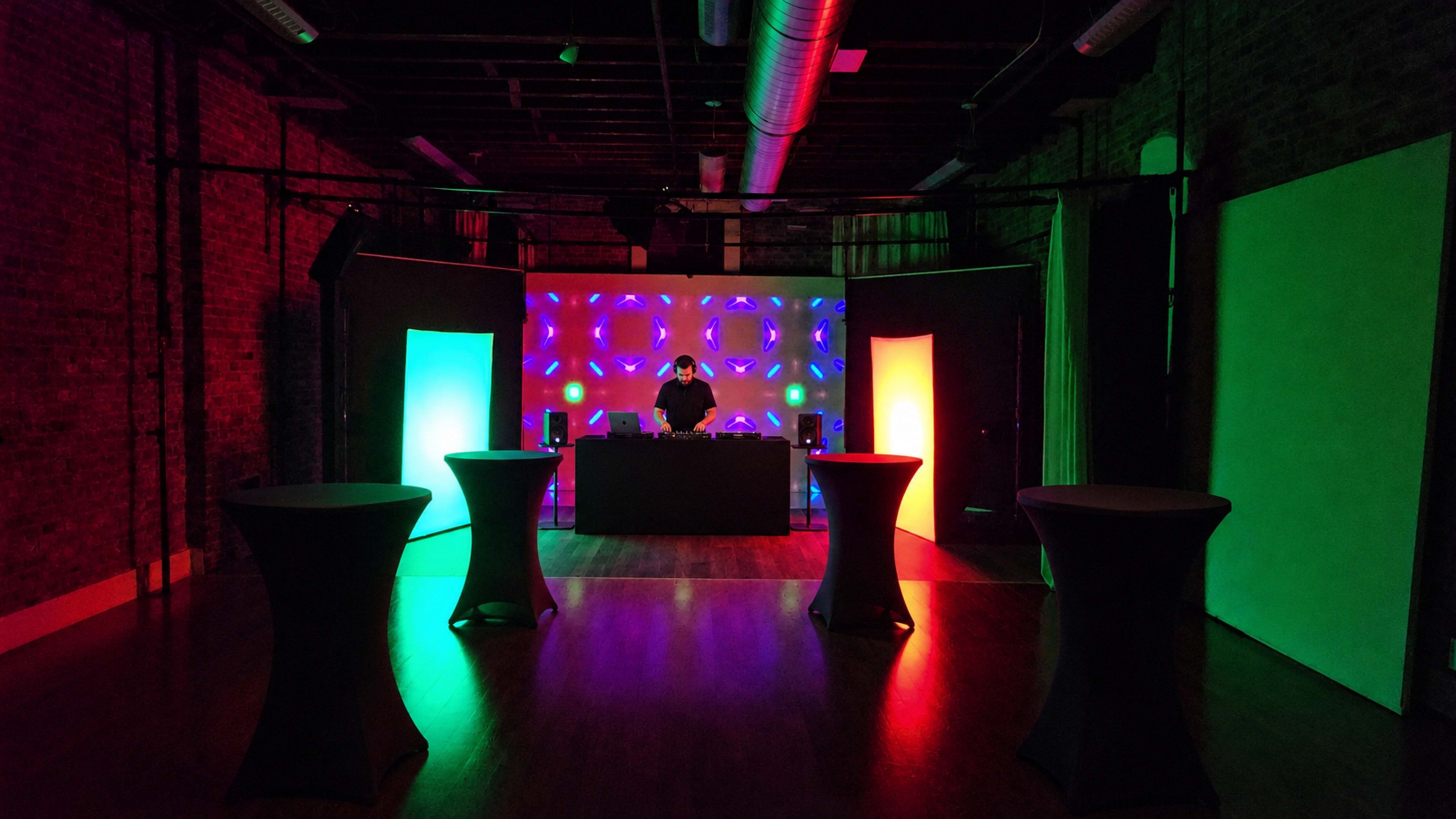 A DJ stands behind a table in a dimly lit venue with colorful lights illuminating the space and black tables arranged around him.
