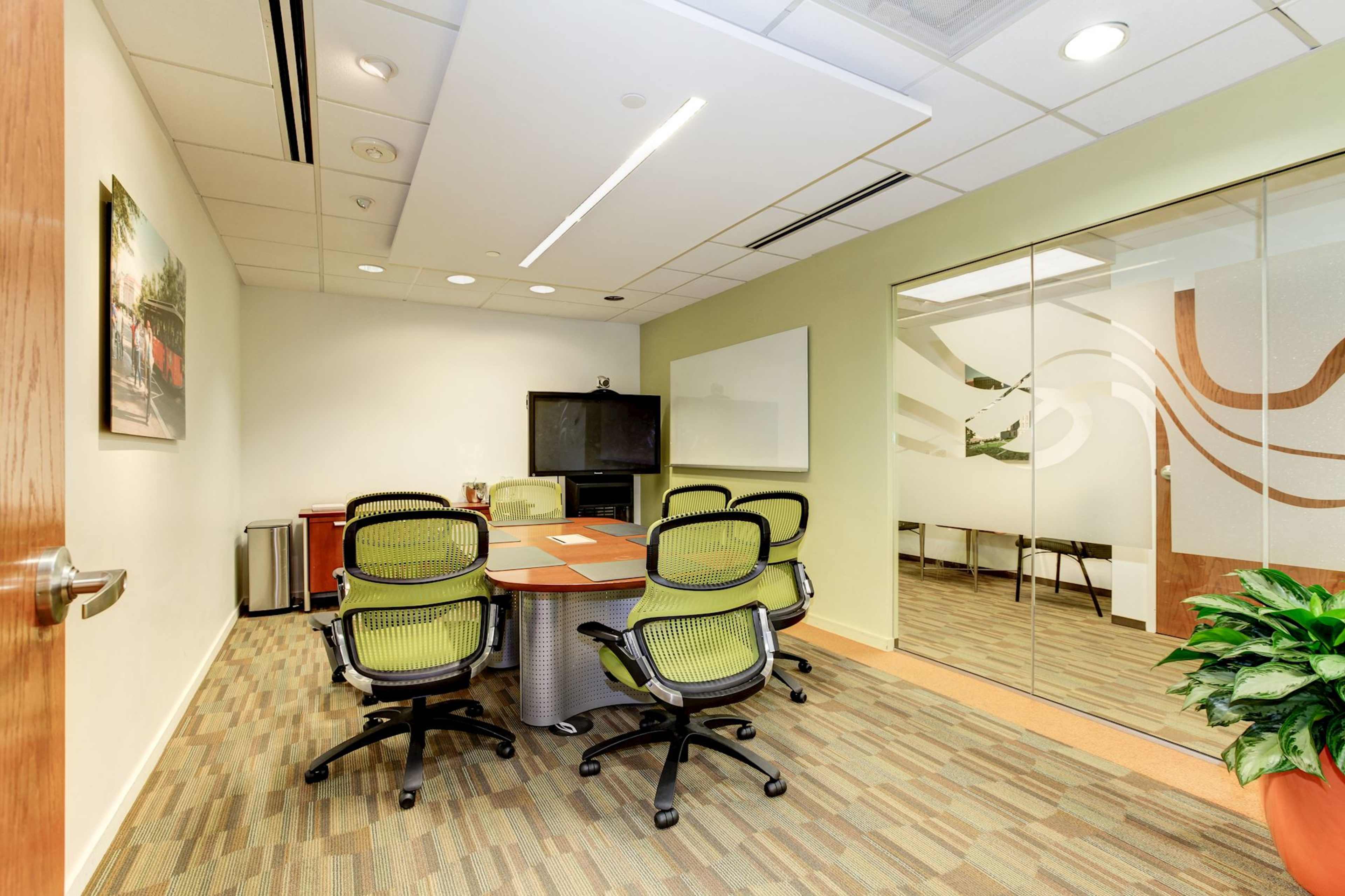 A modern conference room features a round table surrounded by green office chairs, with a television and whiteboard visible on the walls.