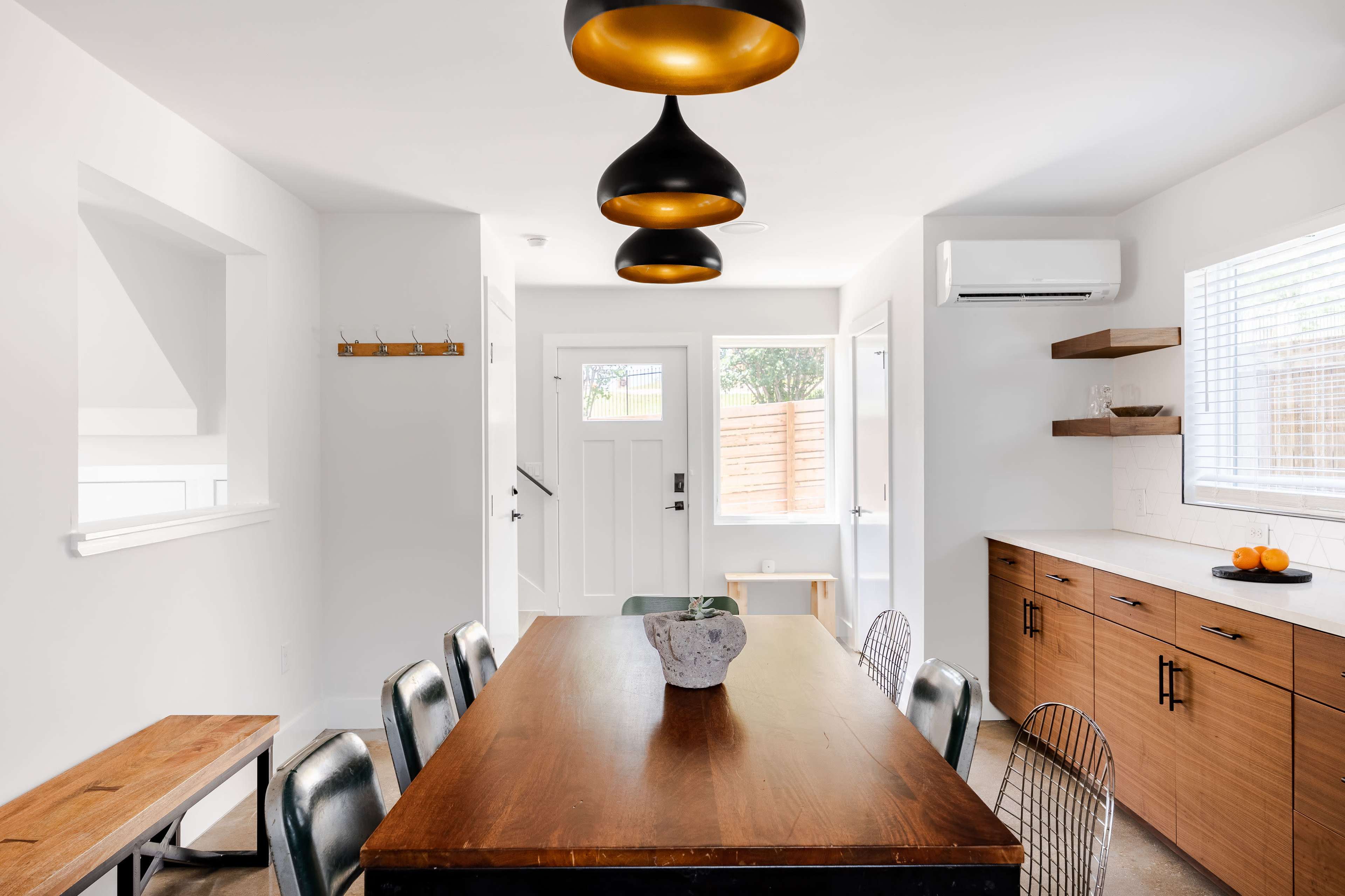 A modern dining area features a wooden table surrounded by metal chairs, illuminated by pendant lights, with a view of a door and window leading to an outdoor space.