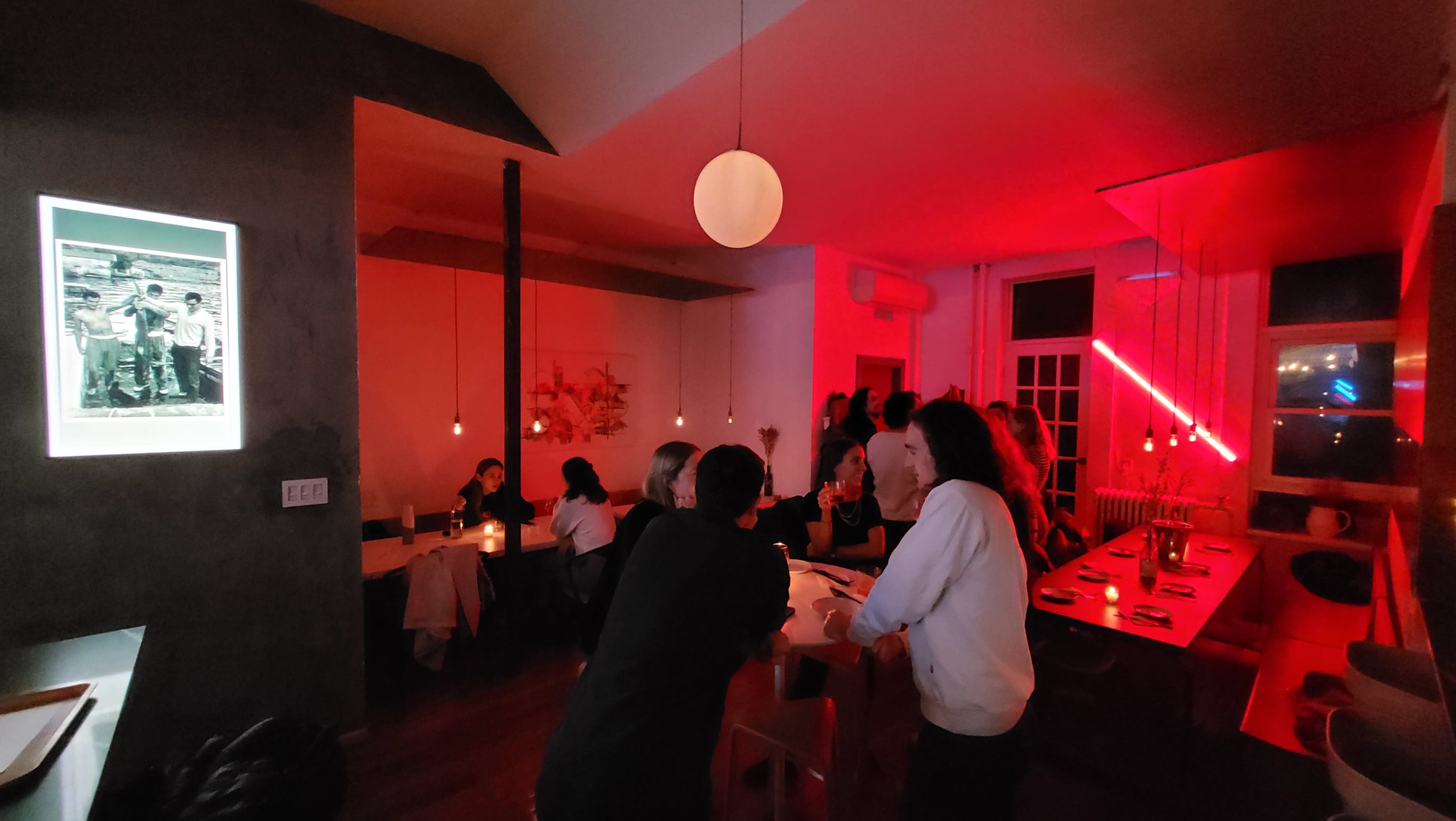 The scene depicts a lively restaurant with patrons gathered at the bar and tables, illuminated by soft red lighting and pendant lamps.
