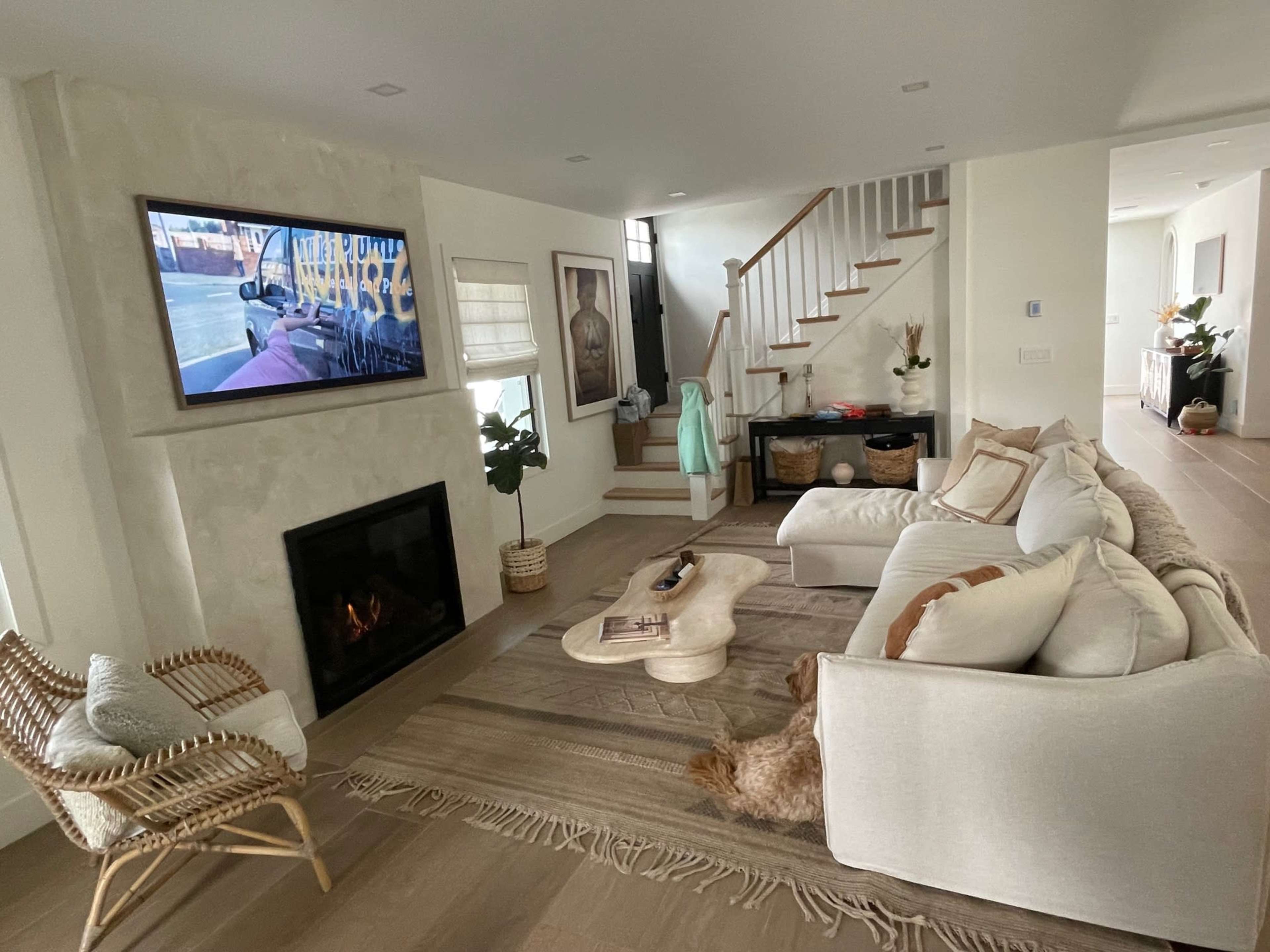 A modern living room features a white sectional sofa, a coffee table, a fireplace, and a television mounted on the wall, with stairs leading to an upper level.