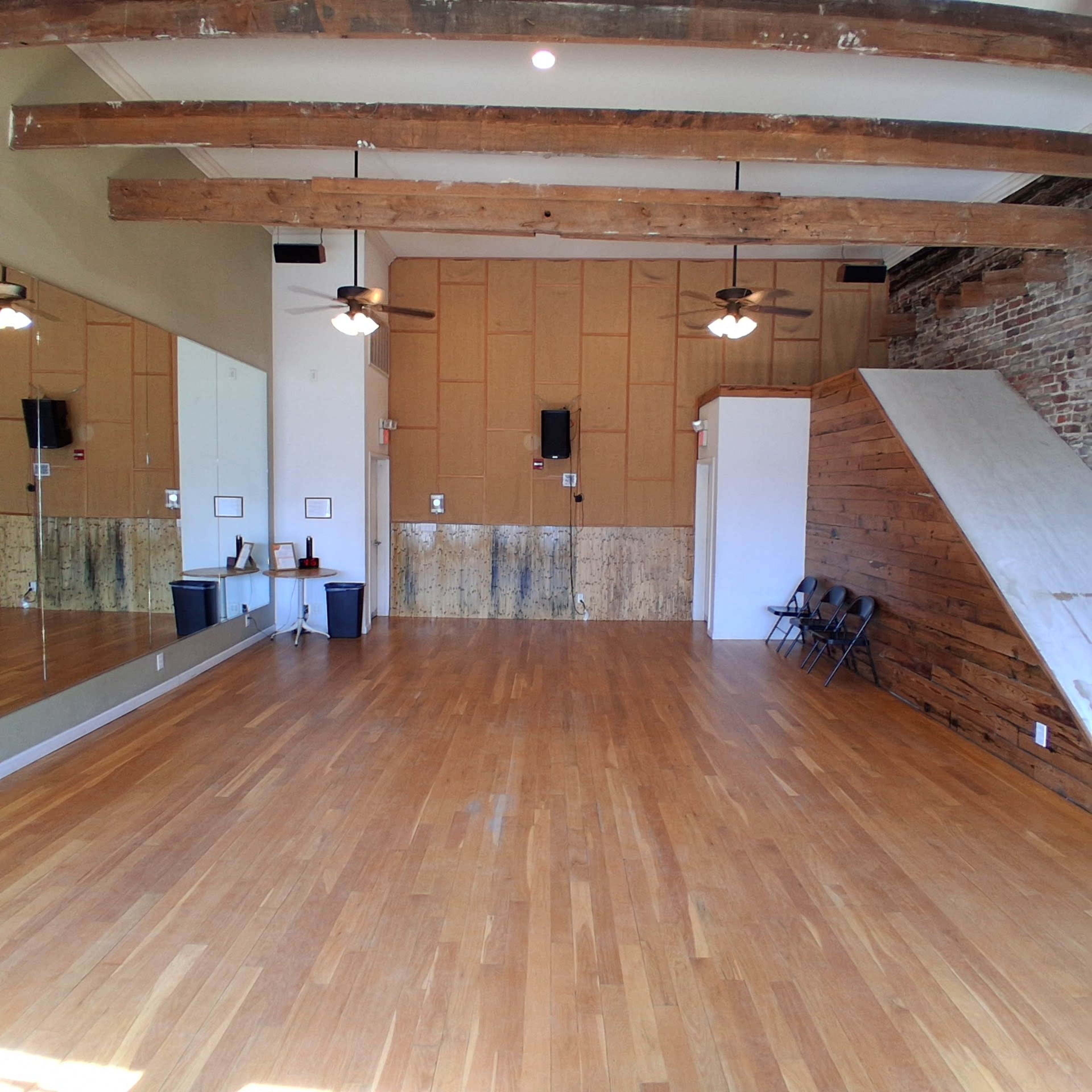 The image shows a spacious dance studio featuring wooden flooring, exposed beams, a mirrored wall, and a small set of black chairs along one side.
